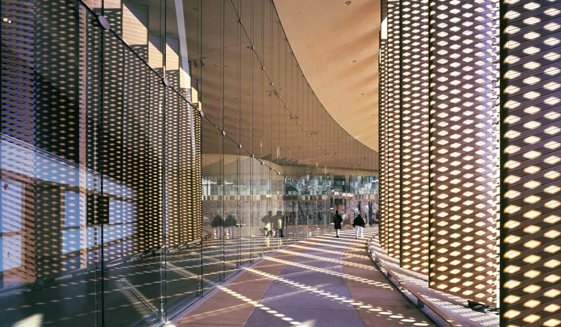 A modern corridor with perforated metal mesh walls illuminated by integrated lighting, creating dramatic shadows on the polished floor, designed by Rafael Vinoly Architects.