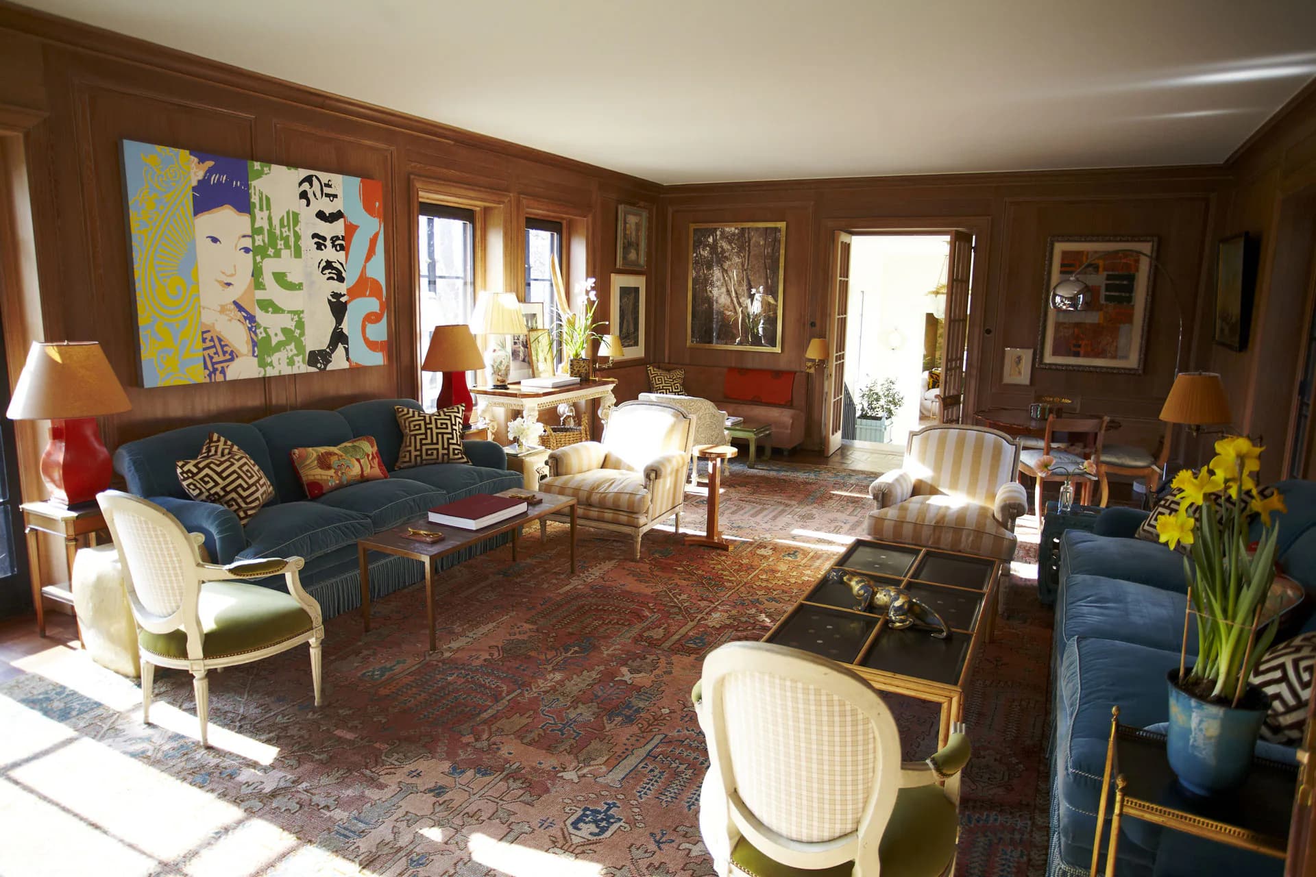 Warm-toned living room featuring blue velvet sofa, cream upholstered chairs, patterned area rug, wooden paneled walls, and colorful artwork, designed by Redd Kaihoi.