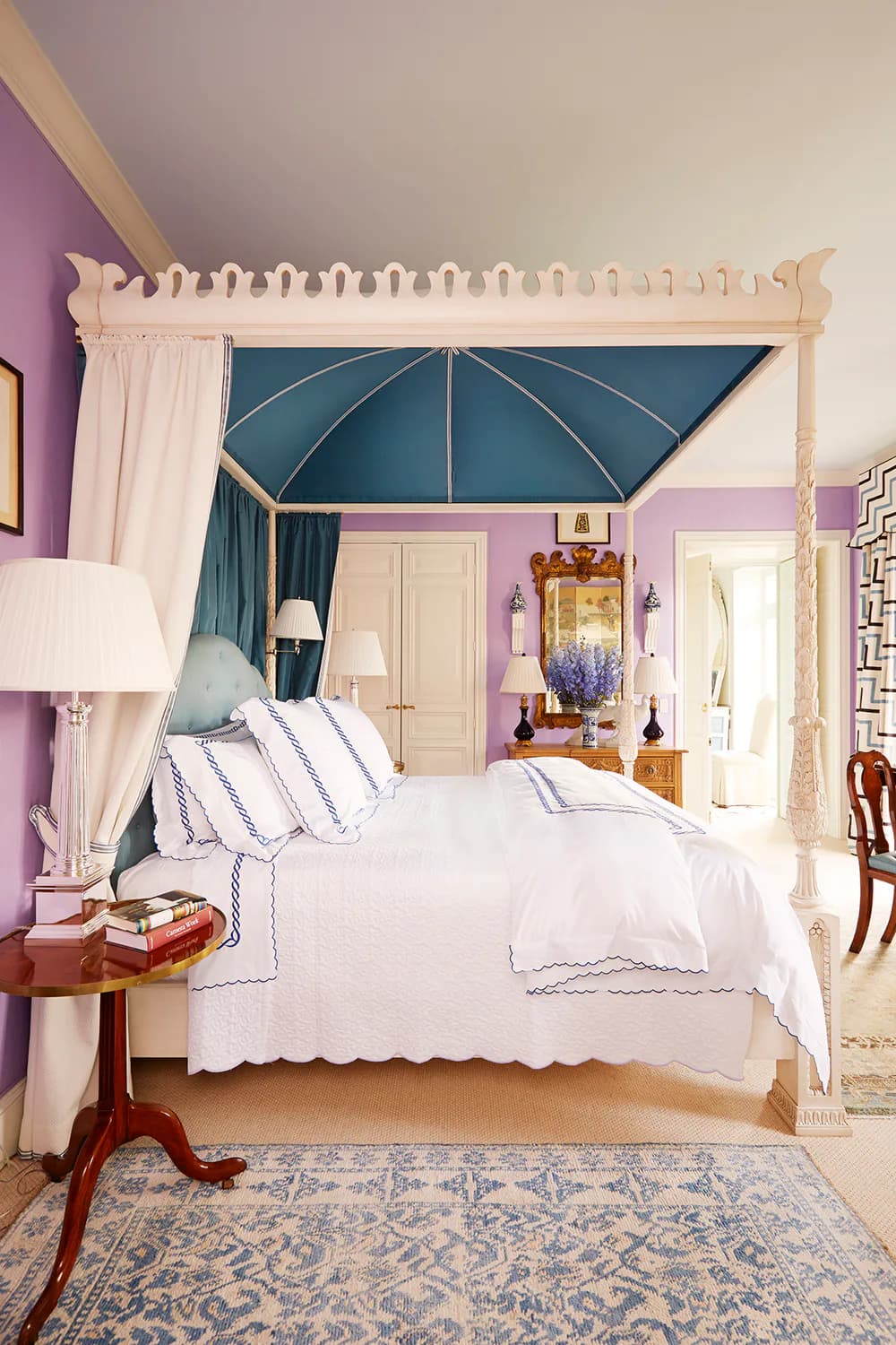 A luxurious bedroom features a cream four-poster canopy bed with blue teal fabric, purple accent walls, white bedding with blue piping, and layered cream curtains, designed by Redd Kaihoi.