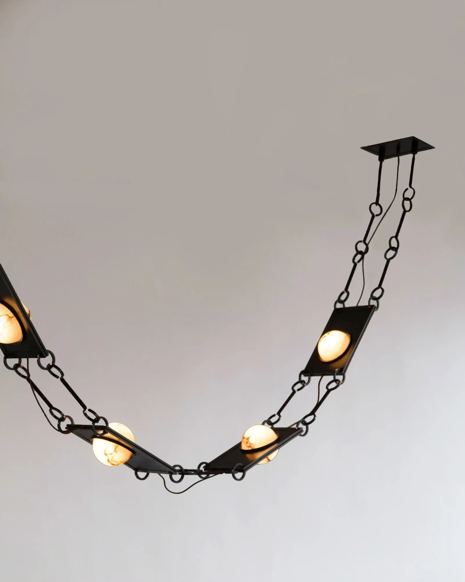 Sculptural pendant lighting installation with curved chain of black metal fixtures and glowing amber bulbs suspended from ceiling mount by Remains Lighting Company.
