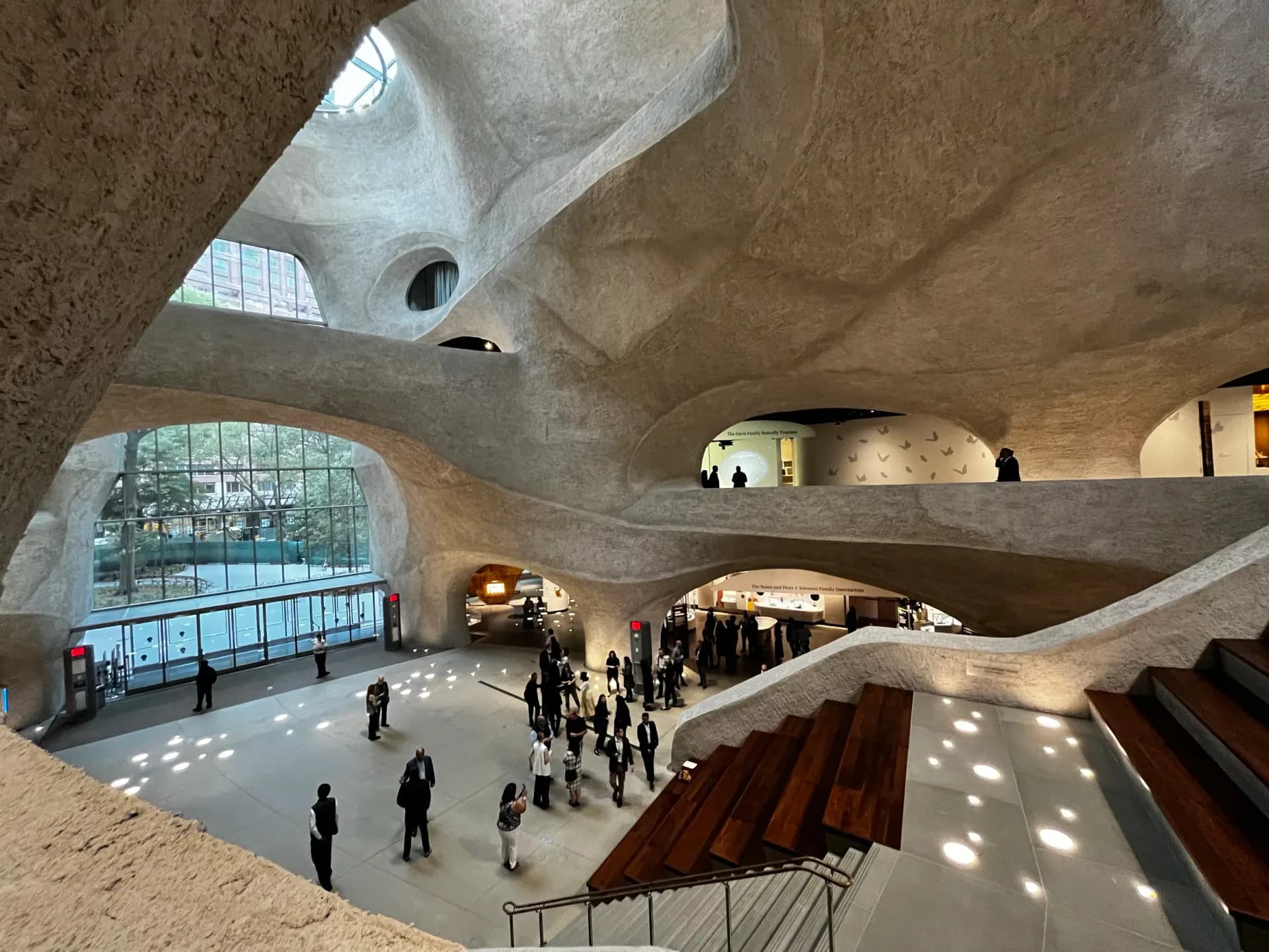 Brutalist concrete interior with organic curved forms, skylights, and grand staircase in warm earth tones by Renfro Design Group.