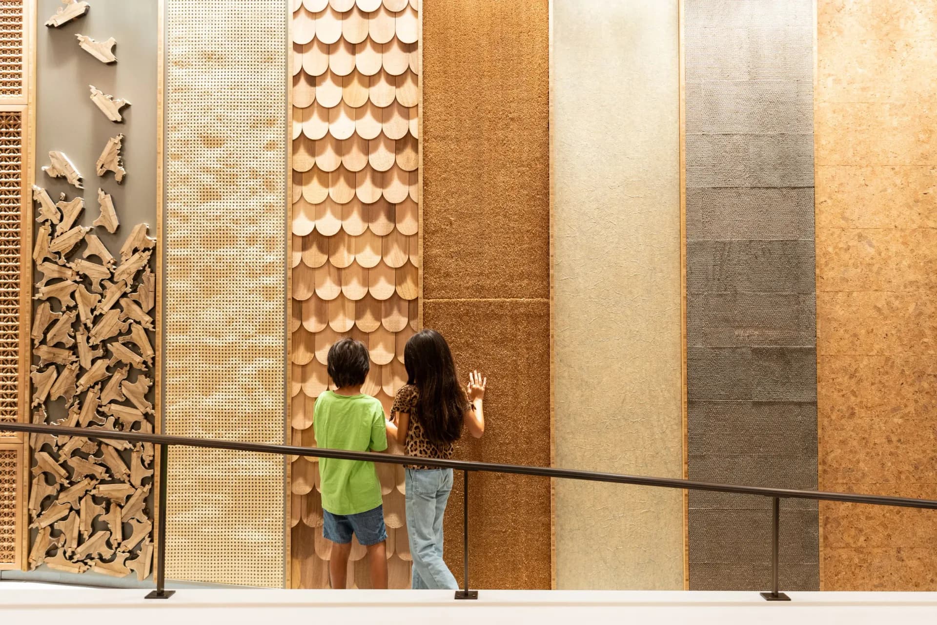 Two children view a materials showroom display wall featuring varied tile, stone, and textured finishes in warm and neutral tones by Renfro Design Group.