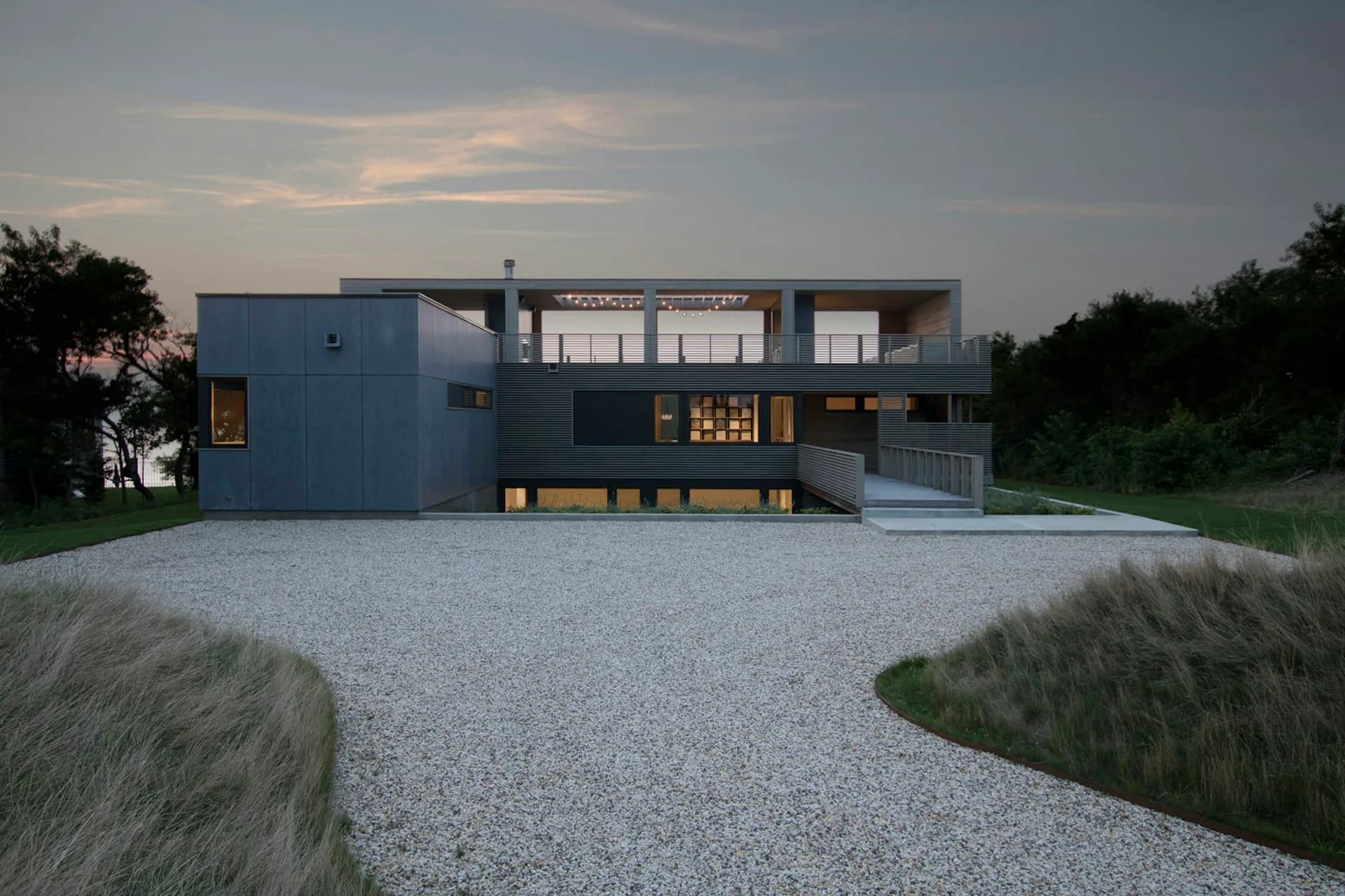 Modern residential architecture featuring a blue-clad cubic form with large windows and a gravel courtyard surrounded by native grasses at dusk by Resolution: 4 Architecture.