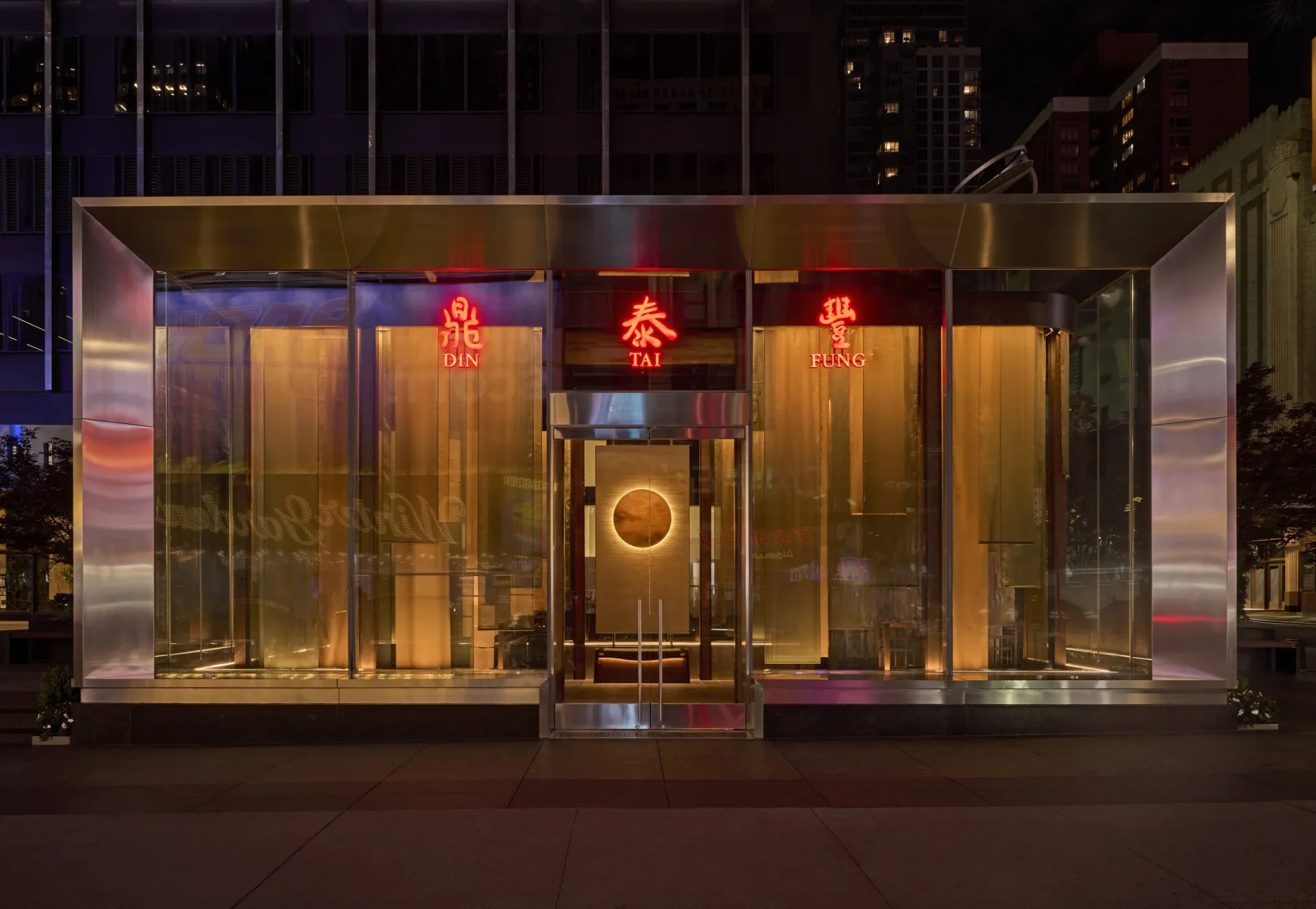 Illuminated storefront with glass facade, warm golden lighting, red neon signage, and metallic framing by Reveal Design Group.