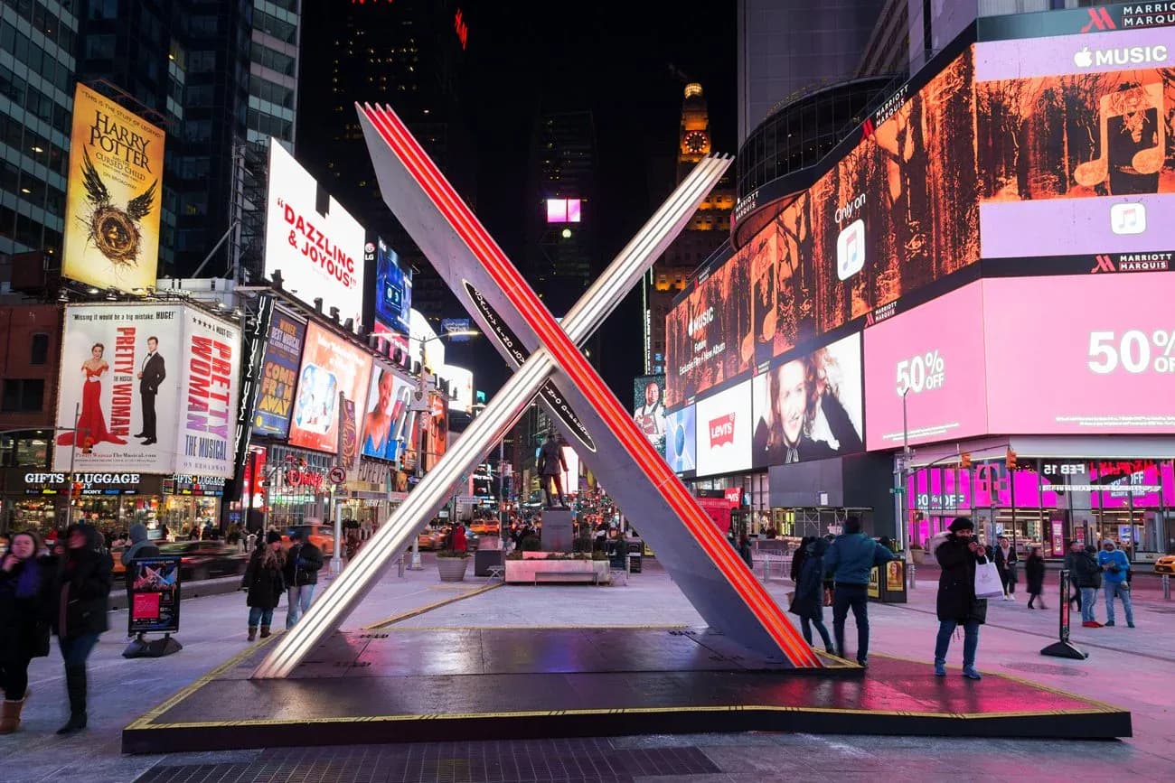 Times Square installation featuring a large white X-shaped sculptural element with red neon accents, surrounded by illuminated digital billboards and pedestrians, by Reveal Design Group.