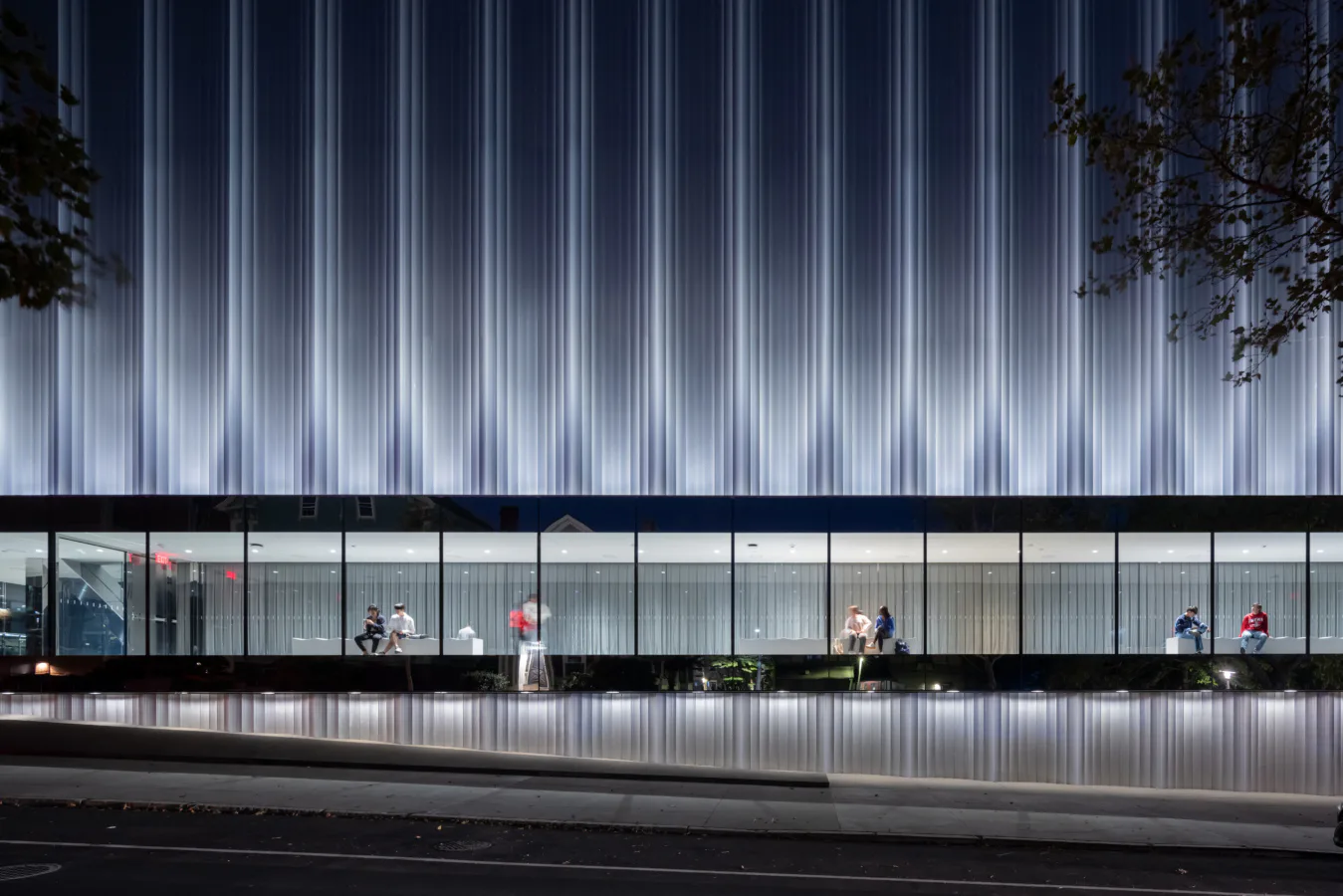 Modern storefront with floor-to-ceiling glass windows, illuminated corrugated metal facade above, and pedestrians visible inside the minimalist retail space by REX.