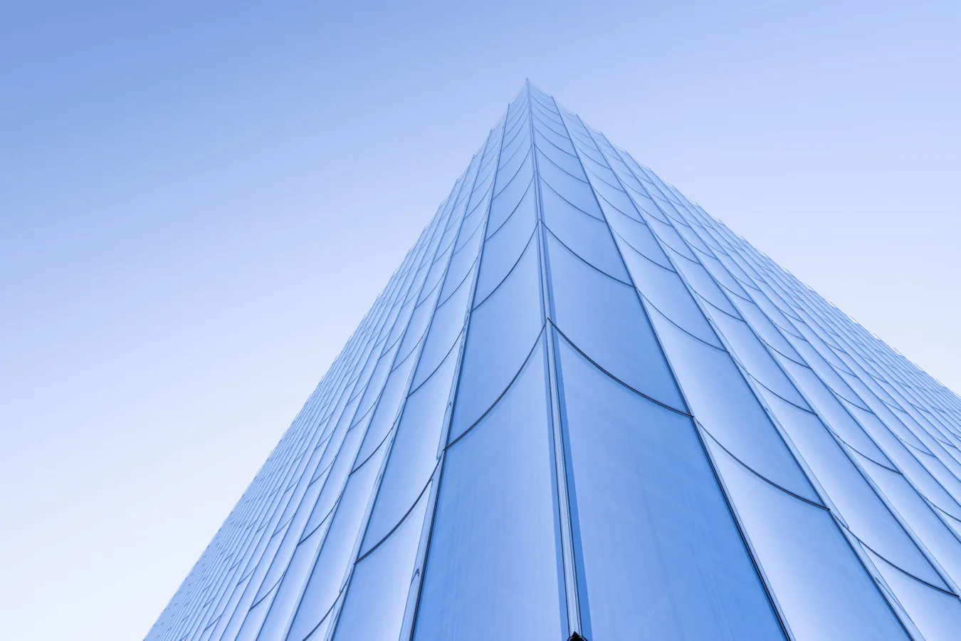 Soaring glass tower facade with geometric curved panels and vertical ribbing ascending to a sharp point against clear sky, by REX.