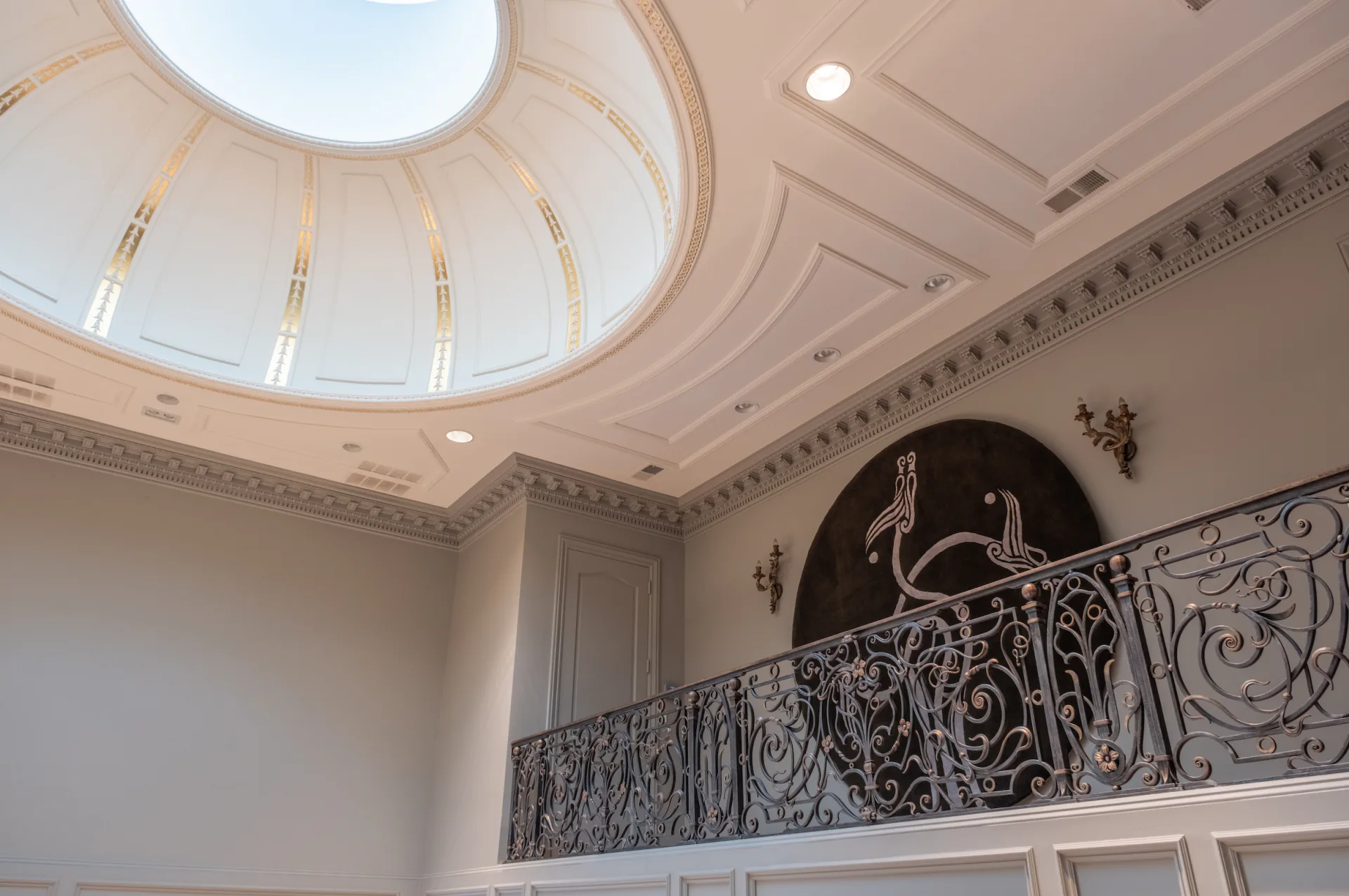 Elegant curved staircase with ornate wrought-iron railing, coffered ceiling with skylight, and classical architectural details by Rhyme Studio.
