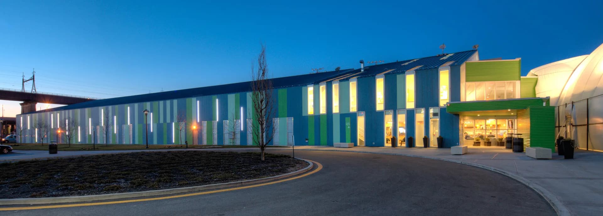 Contemporary institutional building with blue and yellow facade, horizontal window bands, and curved driveway approach at dusk by Ricardo Zurita Architecture and Planning, P.C. (RZAPS).