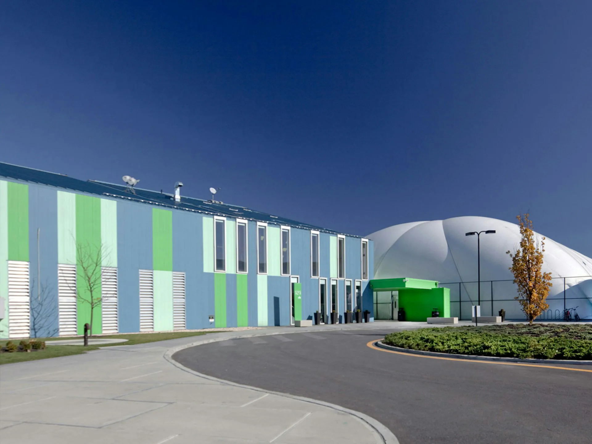 Modern industrial facility with green and white striped facade, curved white building element, and landscaped grounds under clear blue sky by Ricardo Zurita Architecture and Planning, P.C. (RZAPS).