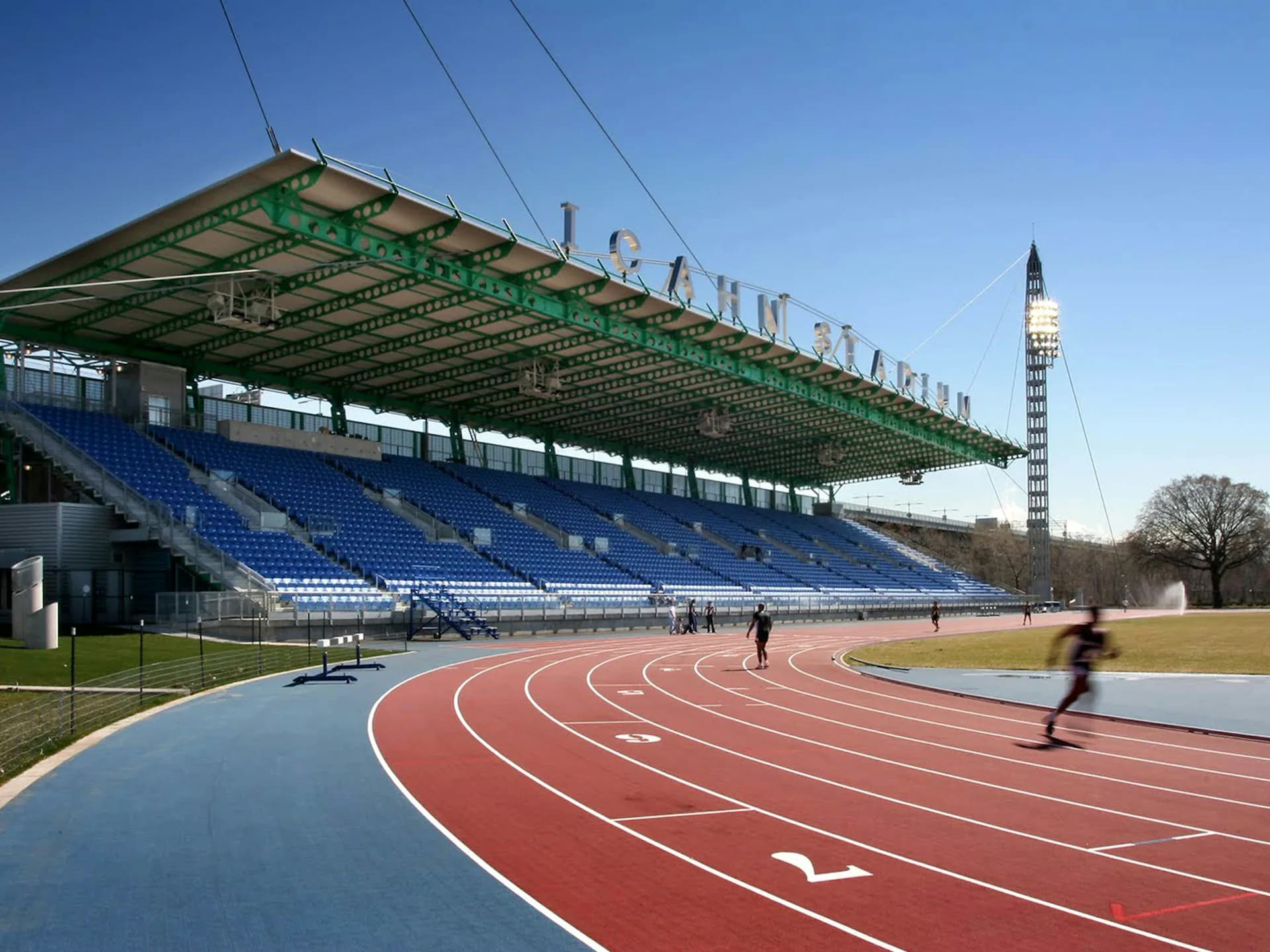 Modern athletic stadium with blue seating, red running track, green roof structure, and tall clock tower in background by Ricardo Zurita Architecture and Planning, P.C. (RZAPS).