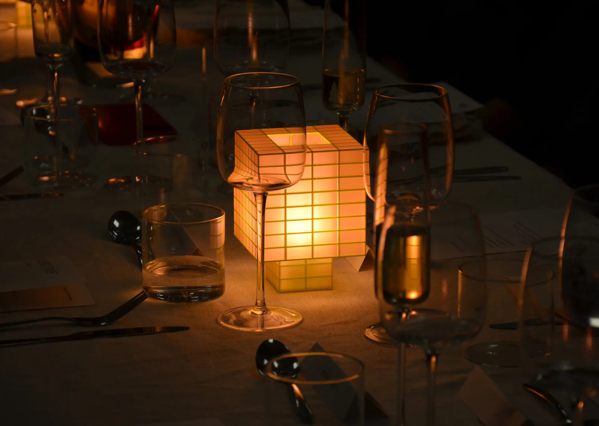 Warm-lit installation featuring a glowing cubic lantern surrounded by glasses and bottles on a reflective dark surface, creating an intimate ambiance by Ridezign.