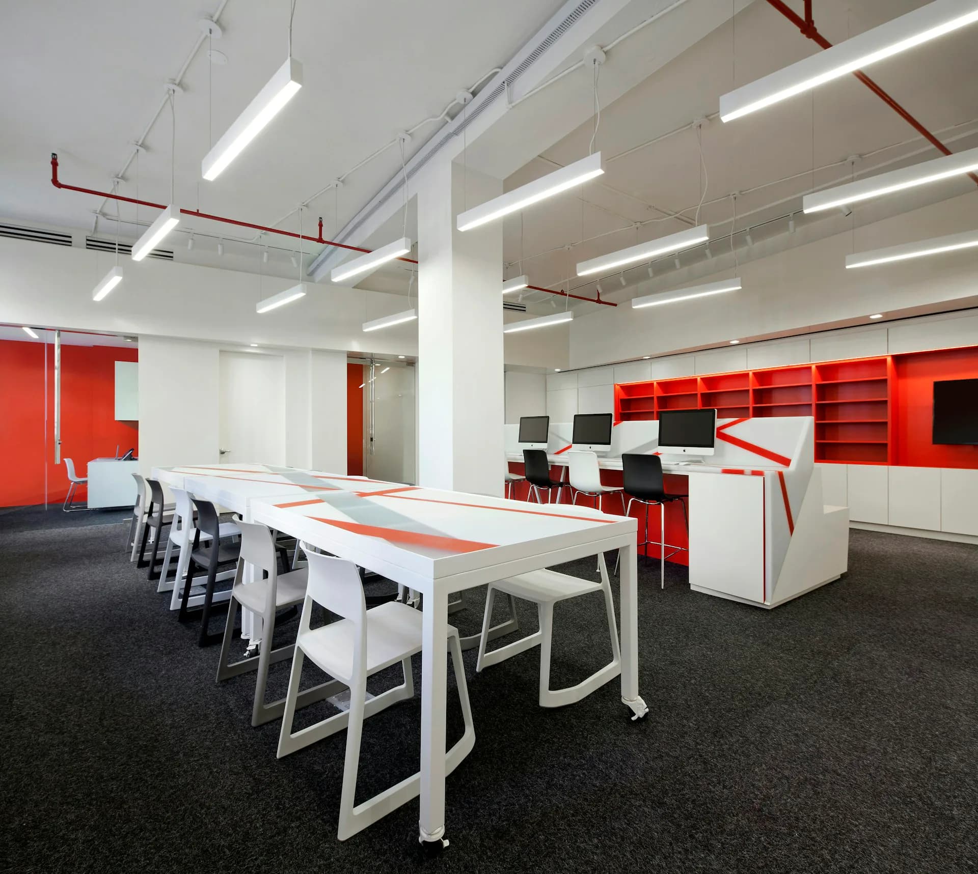 Modern open-plan office space with red and white storage units, white dining tables, and dark flooring, featuring bright LED ceiling lights by Rise Projects.