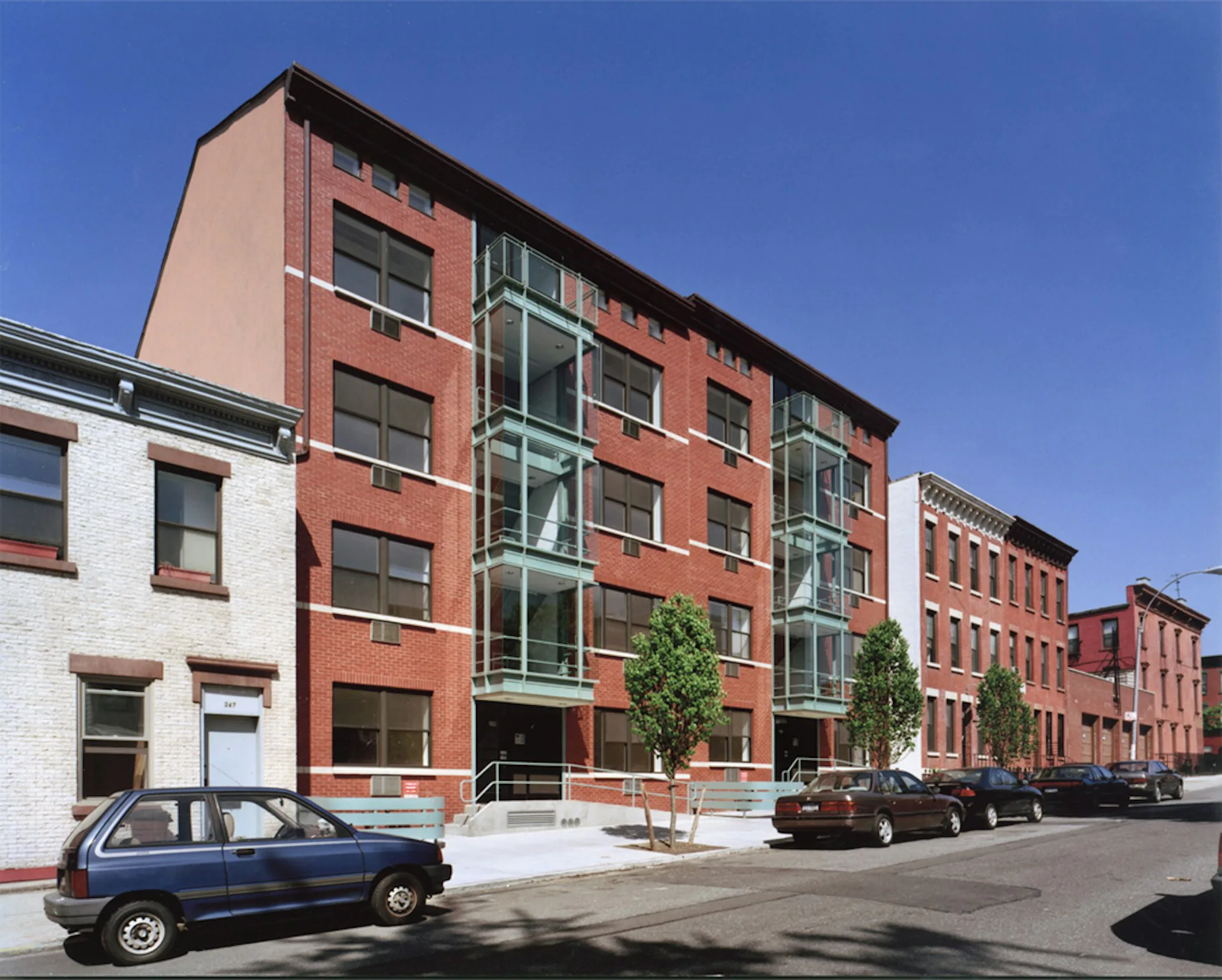Red brick residential building with green metal balconies and ground-floor retail, street view with parked cars by RKTB Architects, P.C.