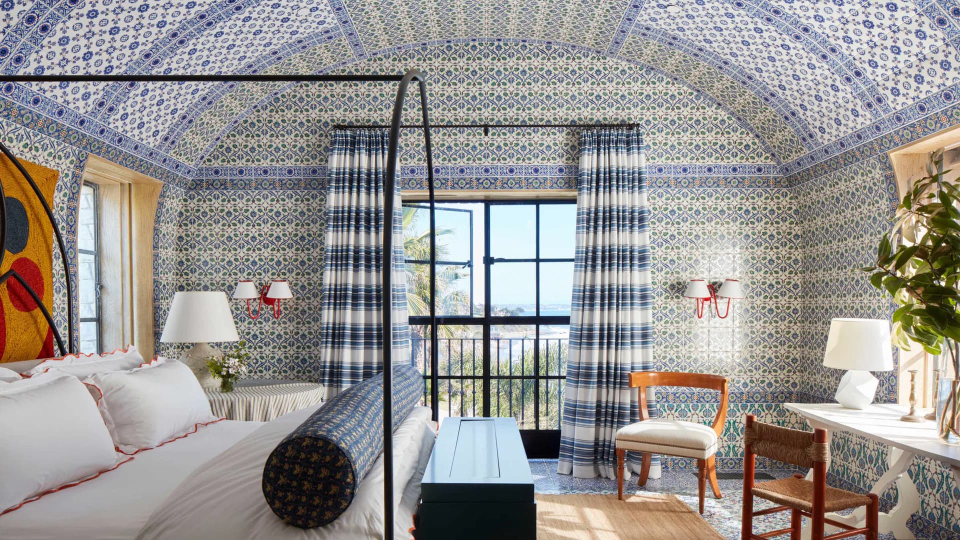 A bedroom with blue geometric patterned wallpaper, black four-poster bed, gingham curtains, and period furnishings by Robert A.M. Stern Architects.