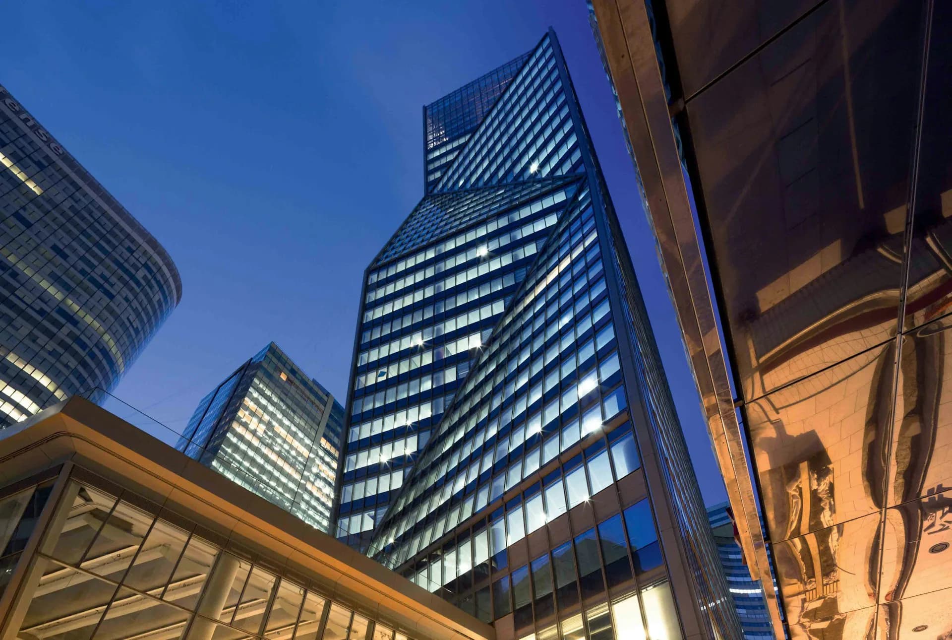 Evening photograph of a tall modern office tower with illuminated diagonal striped facade alongside complementary glass and steel buildings in an urban skyline by Robert A.M. Stern Architects (RAMSA).