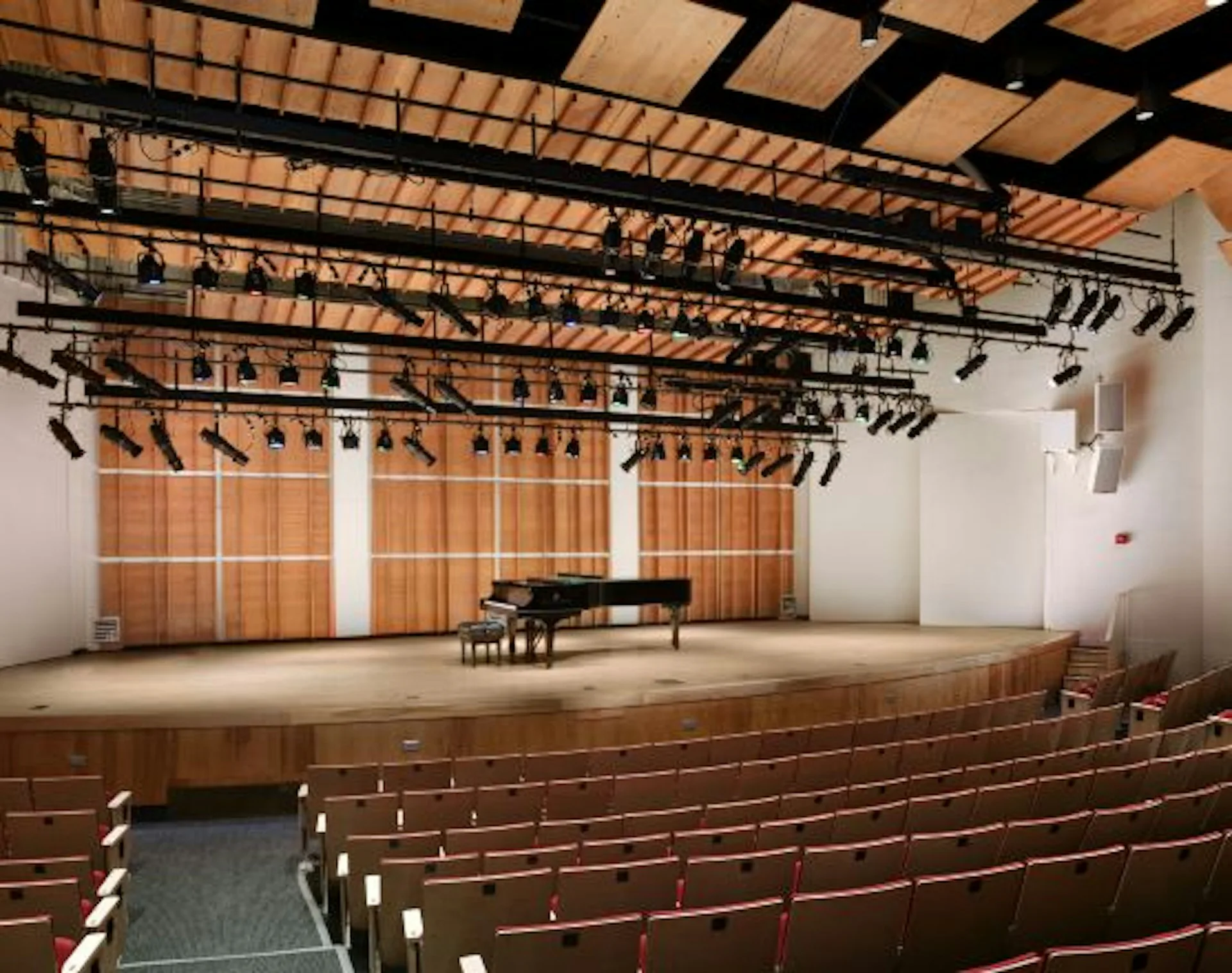 Modern concert hall interior featuring a grand piano on stage, tiered wooden seating, exposed steel truss ceiling with theatrical lighting, and orange-toned wooden wall panels by Robert A.M. Stern Architects (RAMSA).
