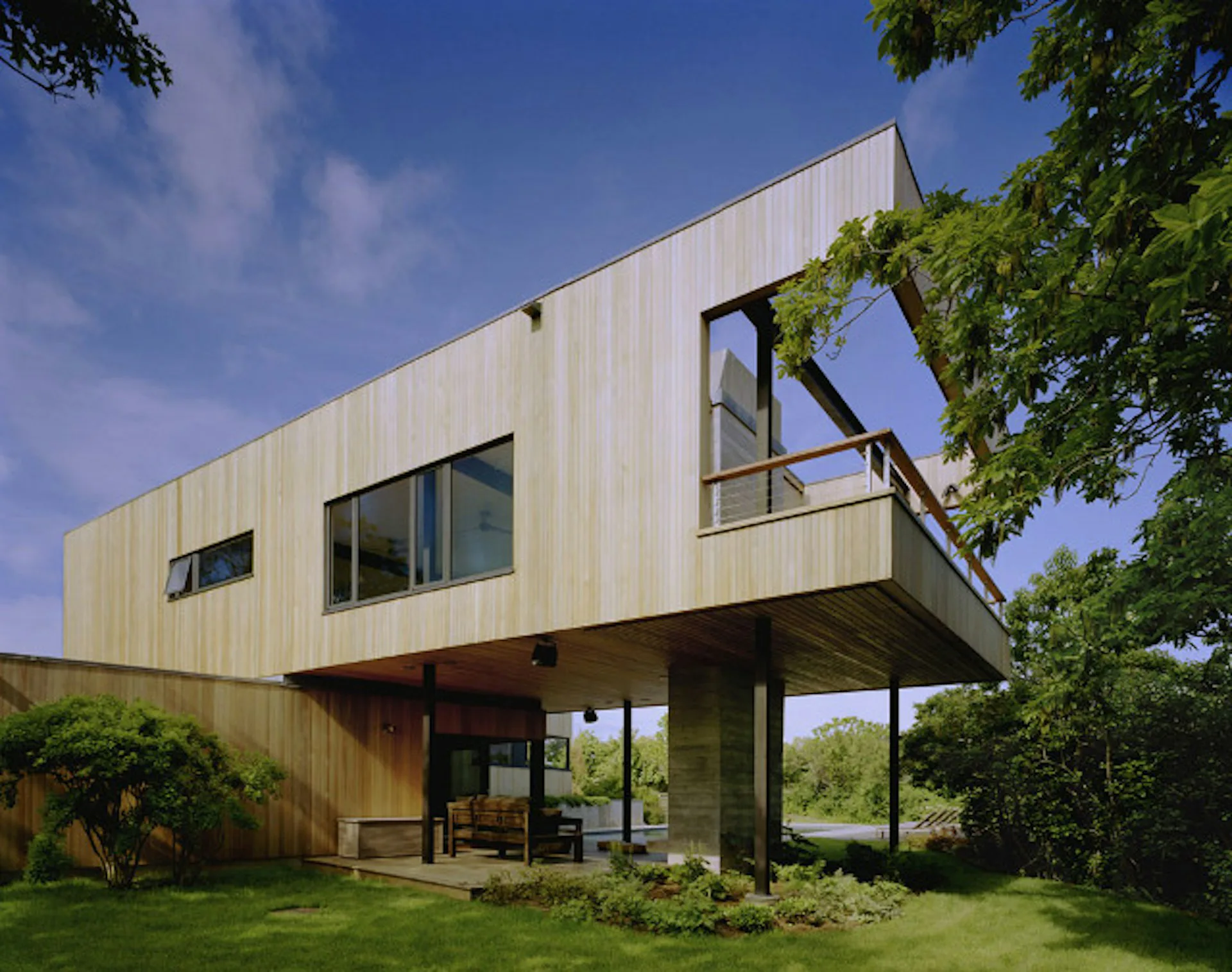 Elevated modernist house with cream-colored metal cladding, concrete pillars, and large glazed openings, surrounded by green lawn and vegetation, by Robert Young Architects.