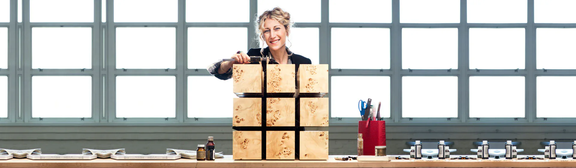 A stylized interior space with large gridded windows features a blonde woman stacking cardboard boxes in a minimalist composition with primary-colored objects and clean geometric forms by RockPaperRobot.