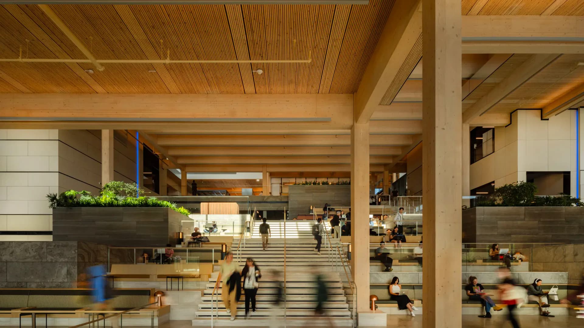 A modern institutional lobby with exposed wooden ceiling beams, stepped seating areas, and blurred figures suggesting movement, designed by Rockwell Group.