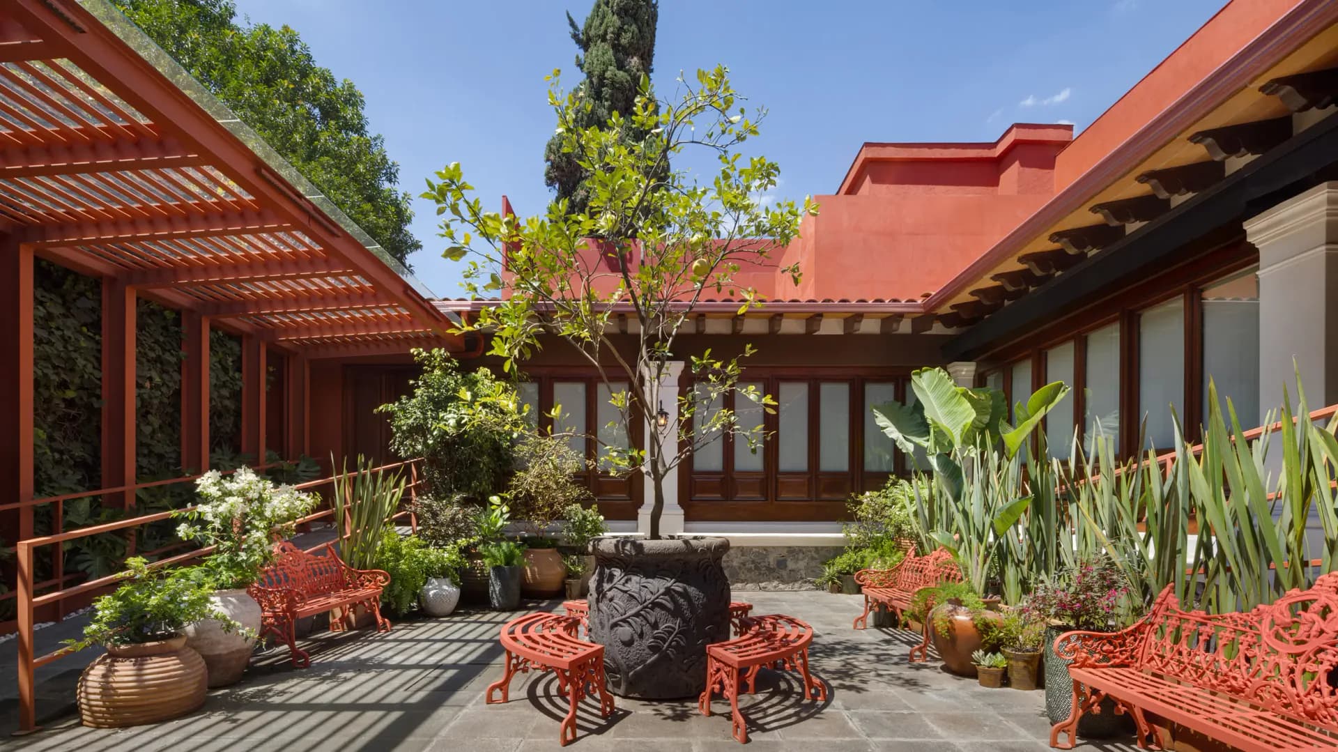 Courtyard with terracotta pergolas, red seating, potted plants, and central tree surrounded by warm-toned architecture by Rockwell Group.
