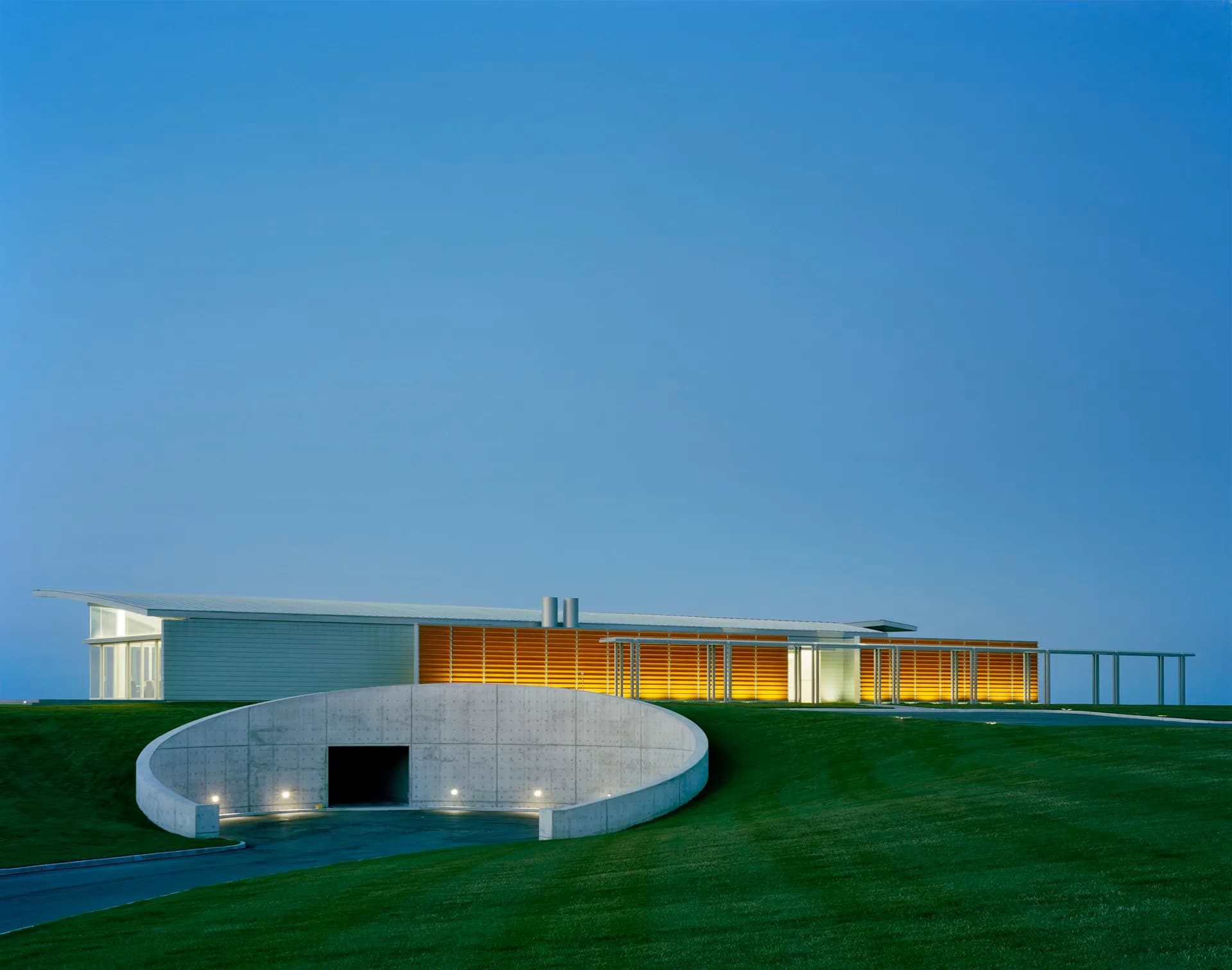 Modern residential structure with curved concrete base, yellow-orange illuminated volumes, and glass walls set on manicured green landscape by Roger Ferris + Partners.