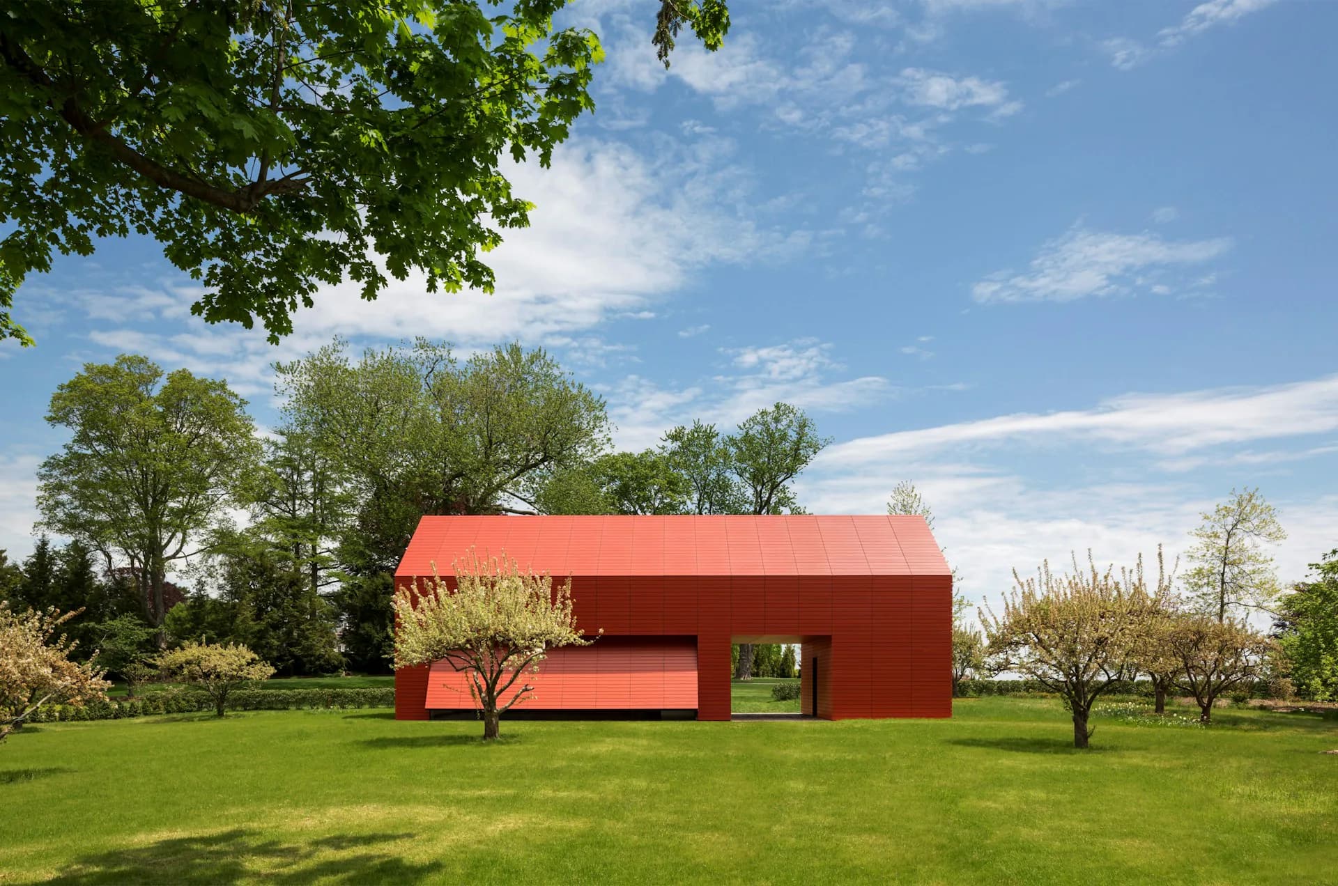 A striking red modern barn structure with clean lines sits in an expansive green meadow surrounded by mature trees, designed by Roger Ferris + Partners.