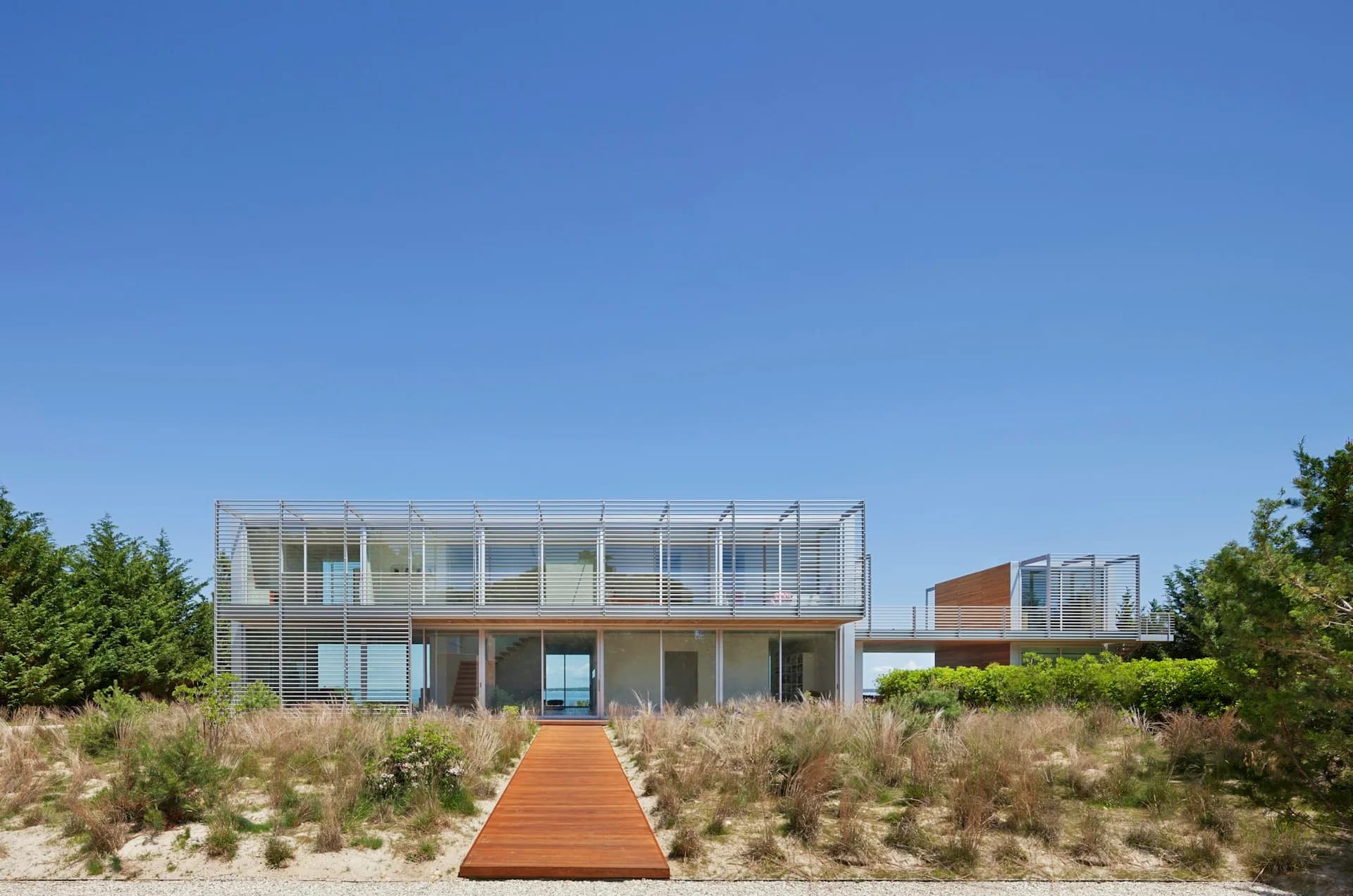 Modern beachfront residence with glass and metal facade, wooden boardwalk entry, and native dune vegetation by Roger Ferris + Partners.