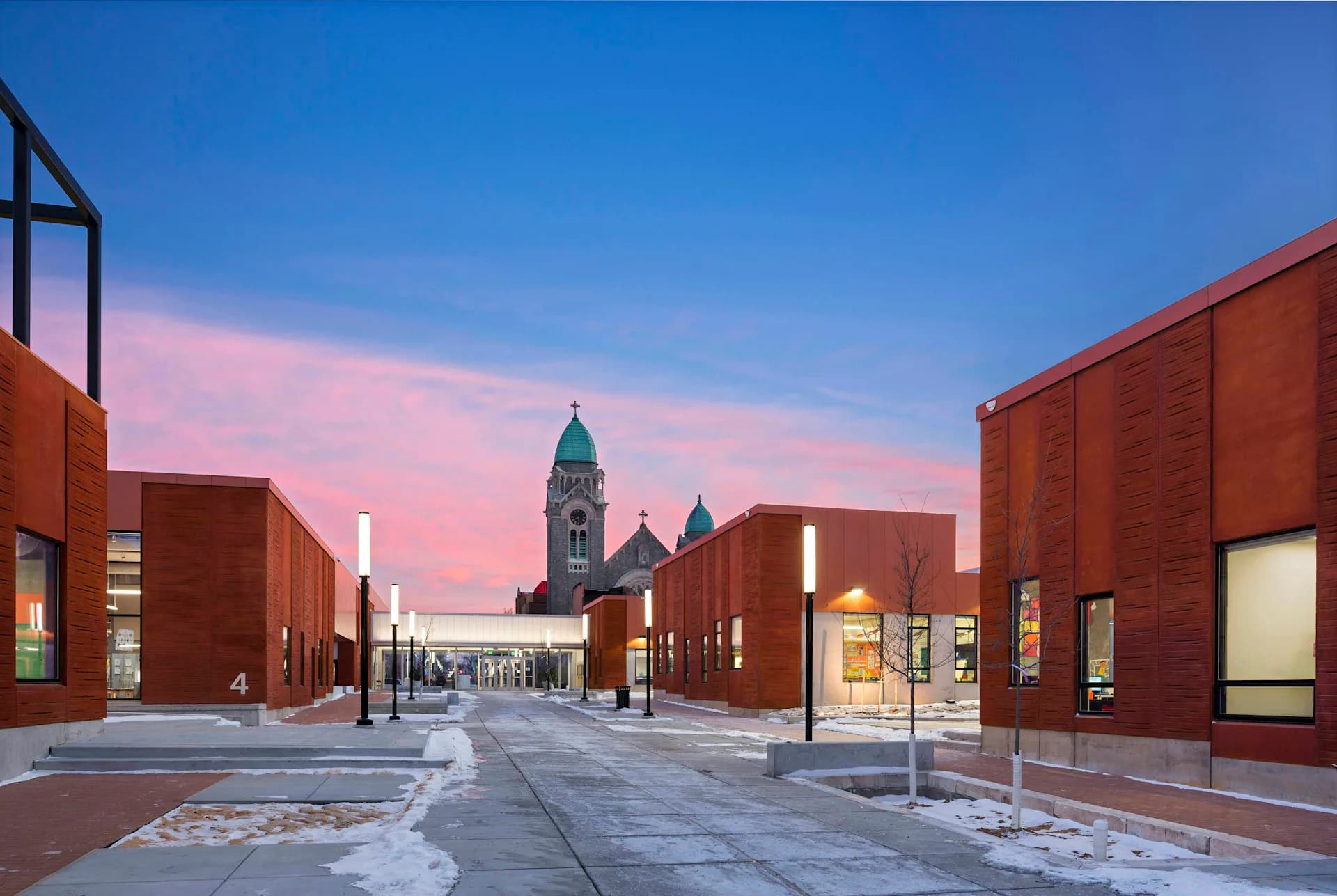 Twilight view of a contemporary red-brick plaza with symmetrical modern buildings framing a street leading to a historic church with spires, designed by ROGERS PARTNERS Architects+Urban Designers.