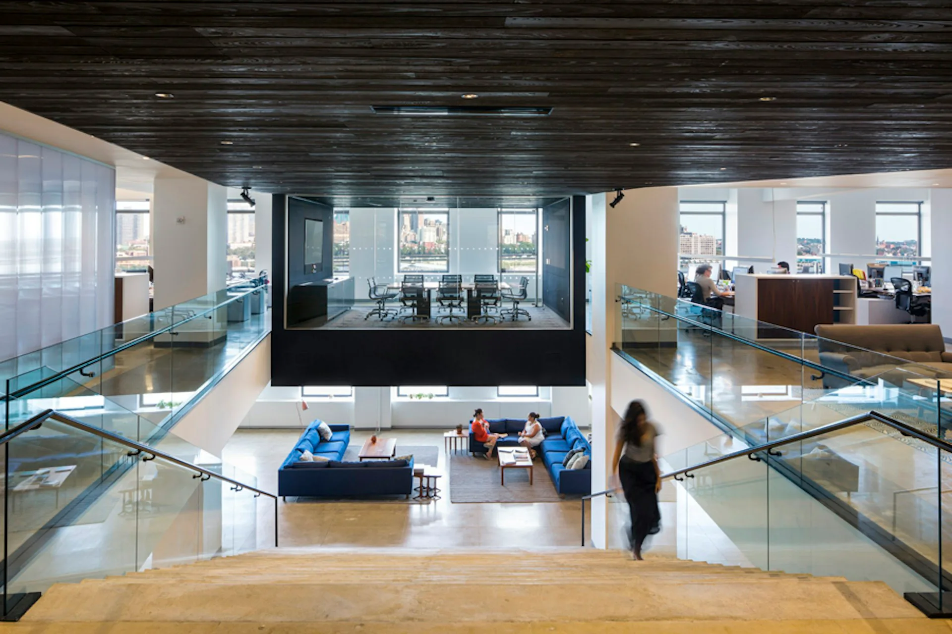 Multi-level open office space with glass partitions, blue furniture clusters, dark ceiling, and natural light from windows, designed by ROGERS PARTNERS Architects+Urban Designers.