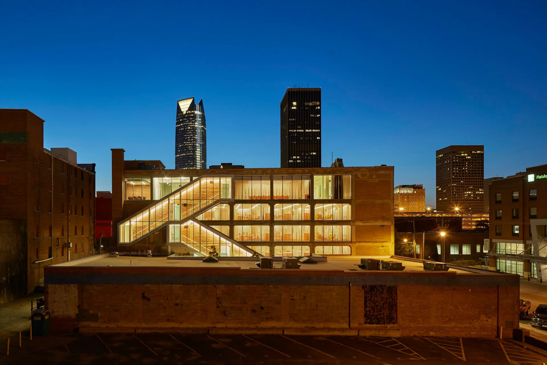 Illuminated modern building with geometric glass facade and warm lighting set against urban skyline at dusk by ROGERS PARTNERS Architects+Urban Designers.