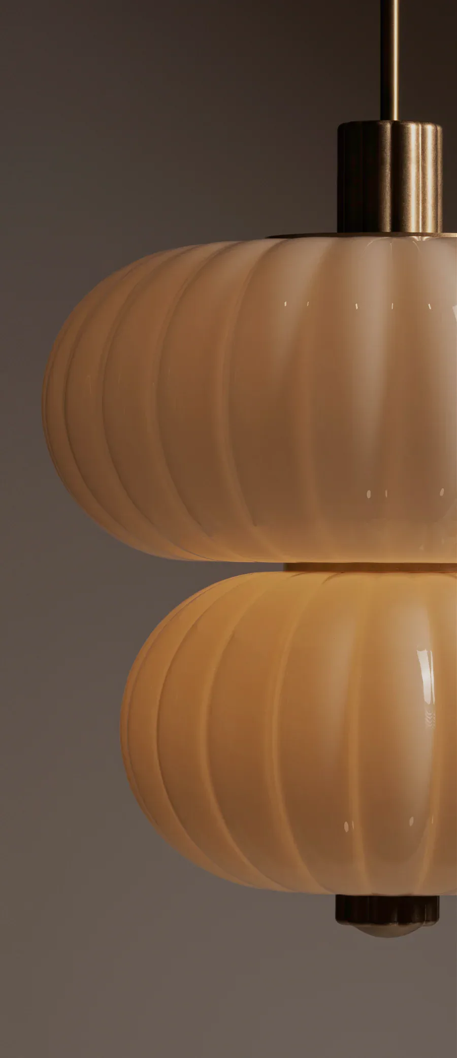 Two stacked pendant lights with ribbed frosted glass shades in warm amber tones and brushed brass fixtures suspended against a neutral gray background by Roll & Hill.