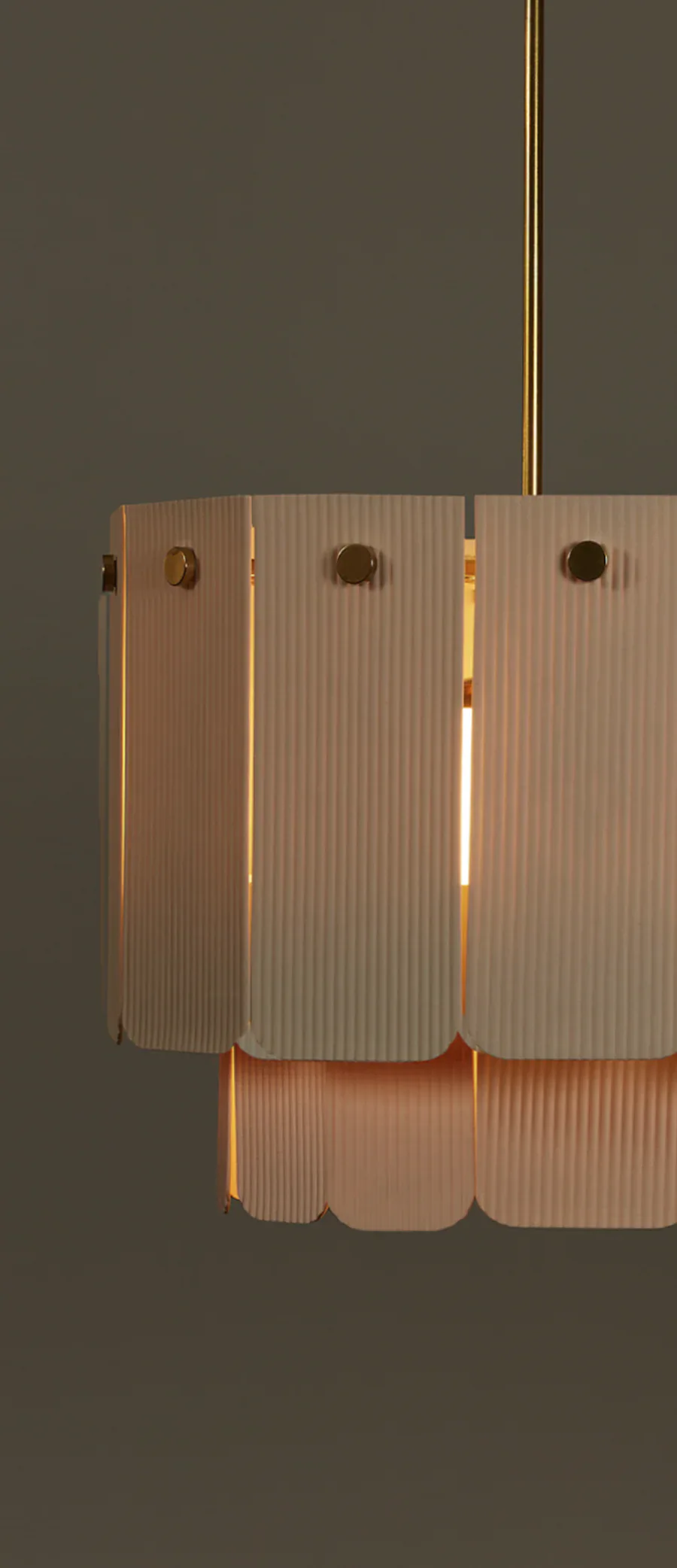 Pendant lighting fixture with ribbed translucent shade panels in warm tones, suspended from brass rod, emitting warm ambient light by Roll & Hill.