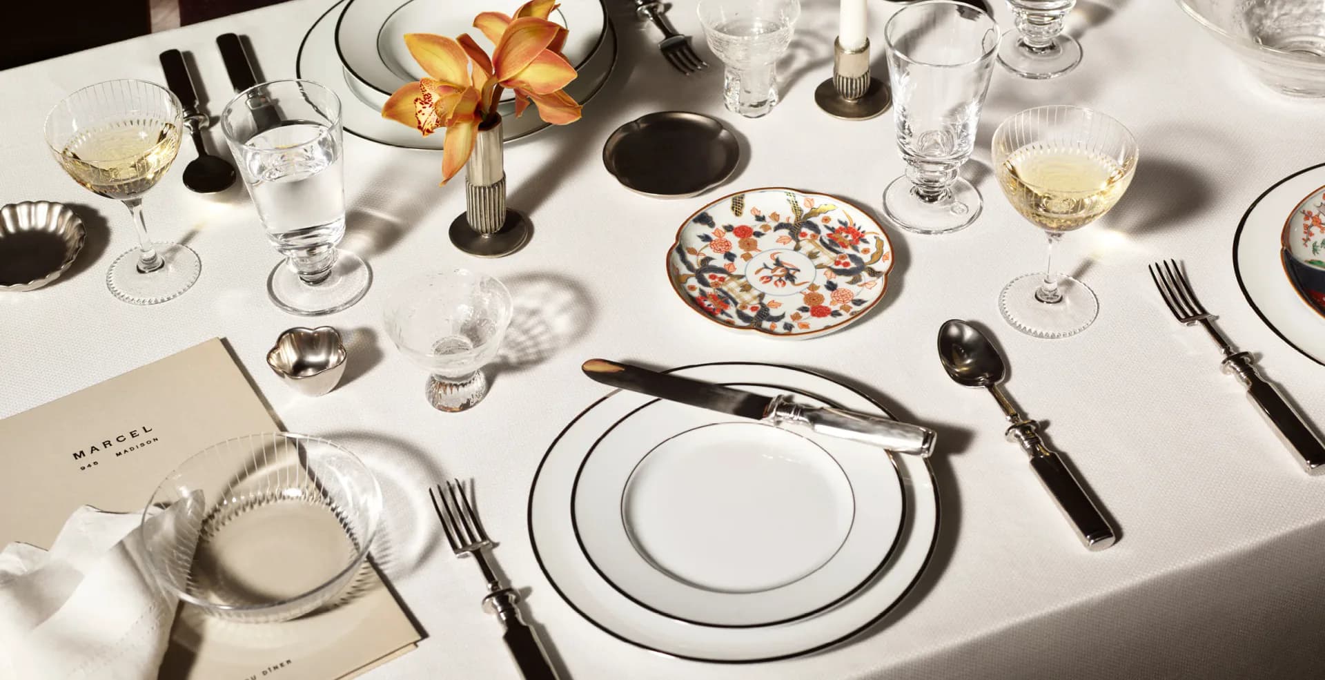 Elegant table setting with white linens, layered plates with graphic borders, black flatware, and gold-accented glassware arranged on a white tablecloth by Roman and Williams.