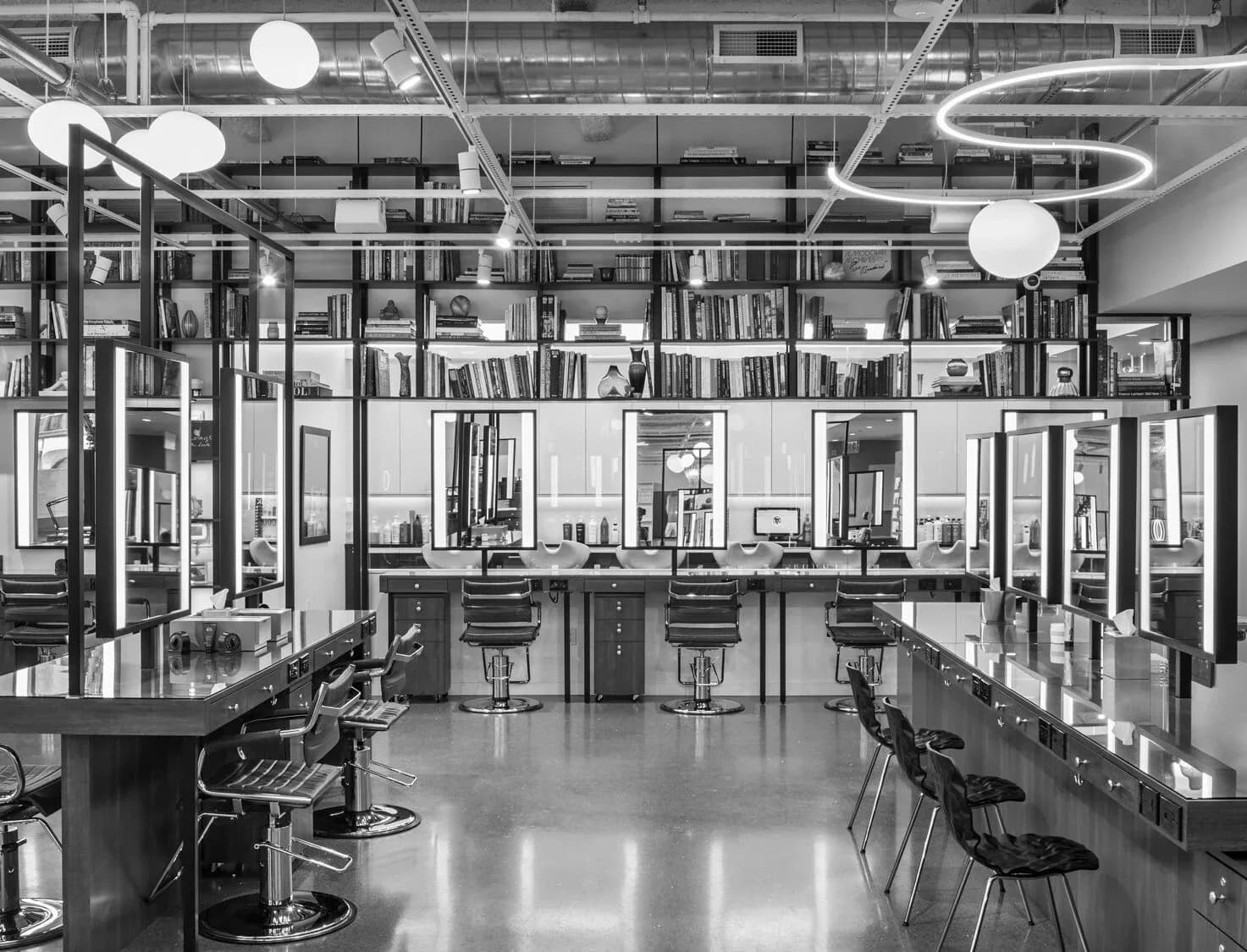Modern salon interior with black and white palette, featuring multiple styling stations with mirrors, shelving for products, polished concrete floors, and contemporary pendant lighting by Ronnette Riley Architect.