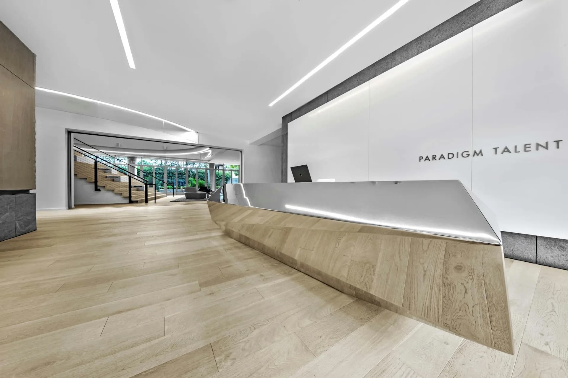 Modern office reception area with curved wooden desk, linear ceiling lights, and Paradigm Talent signage by Rottet Studio.