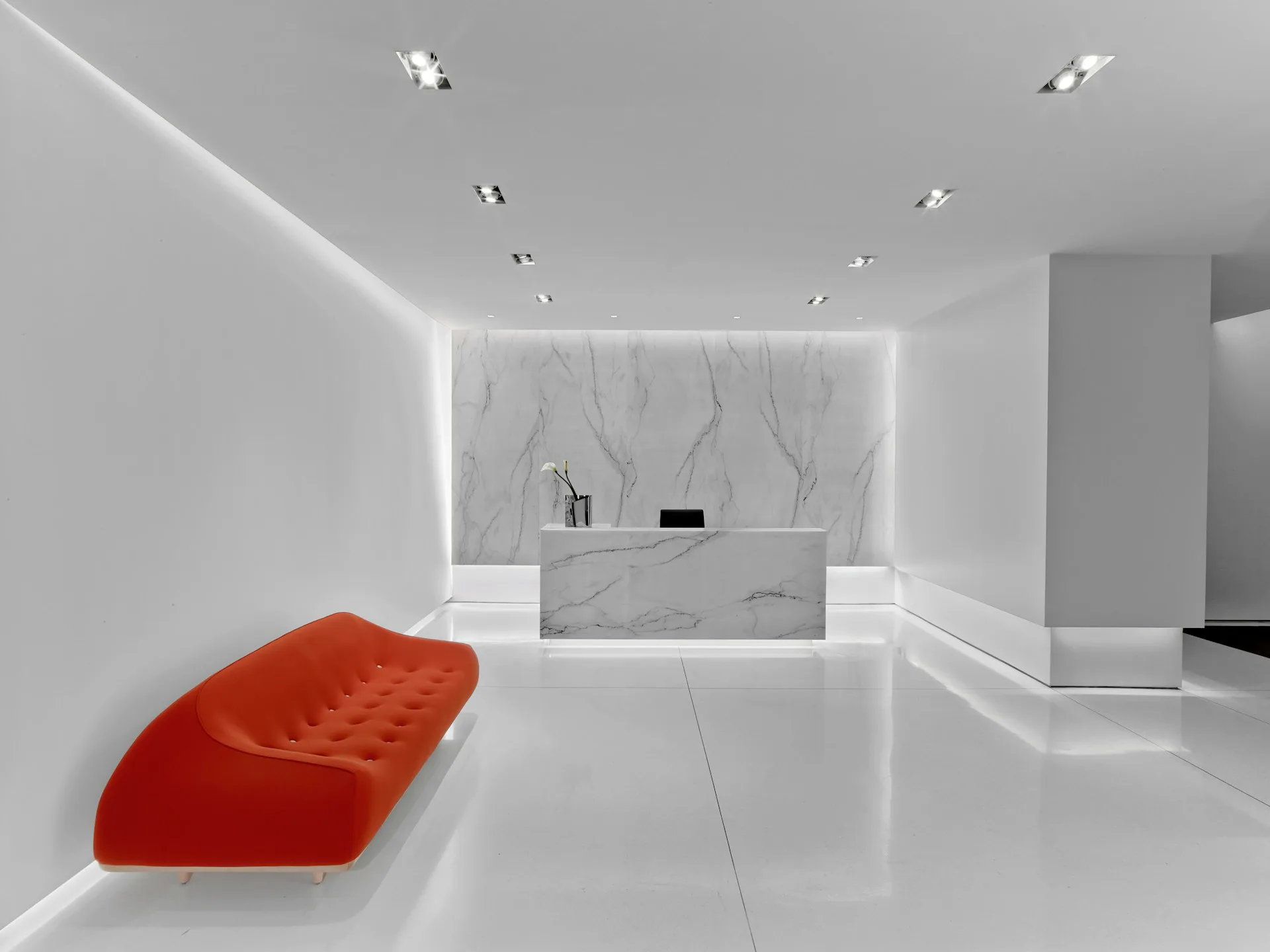 Minimalist showroom interior with white walls, marble display counter, red curved seating element, and recessed ceiling lights by Rottet Studio.