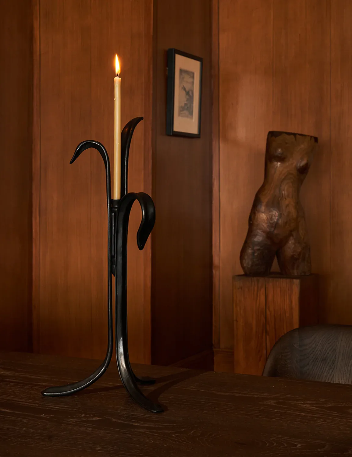 Warm wood-paneled interior featuring a sculptural black iron candelabra with lit candle, wooden torso sculpture on pedestal, and framed artwork, designed by RW Guild (Roman and Williams Guild).