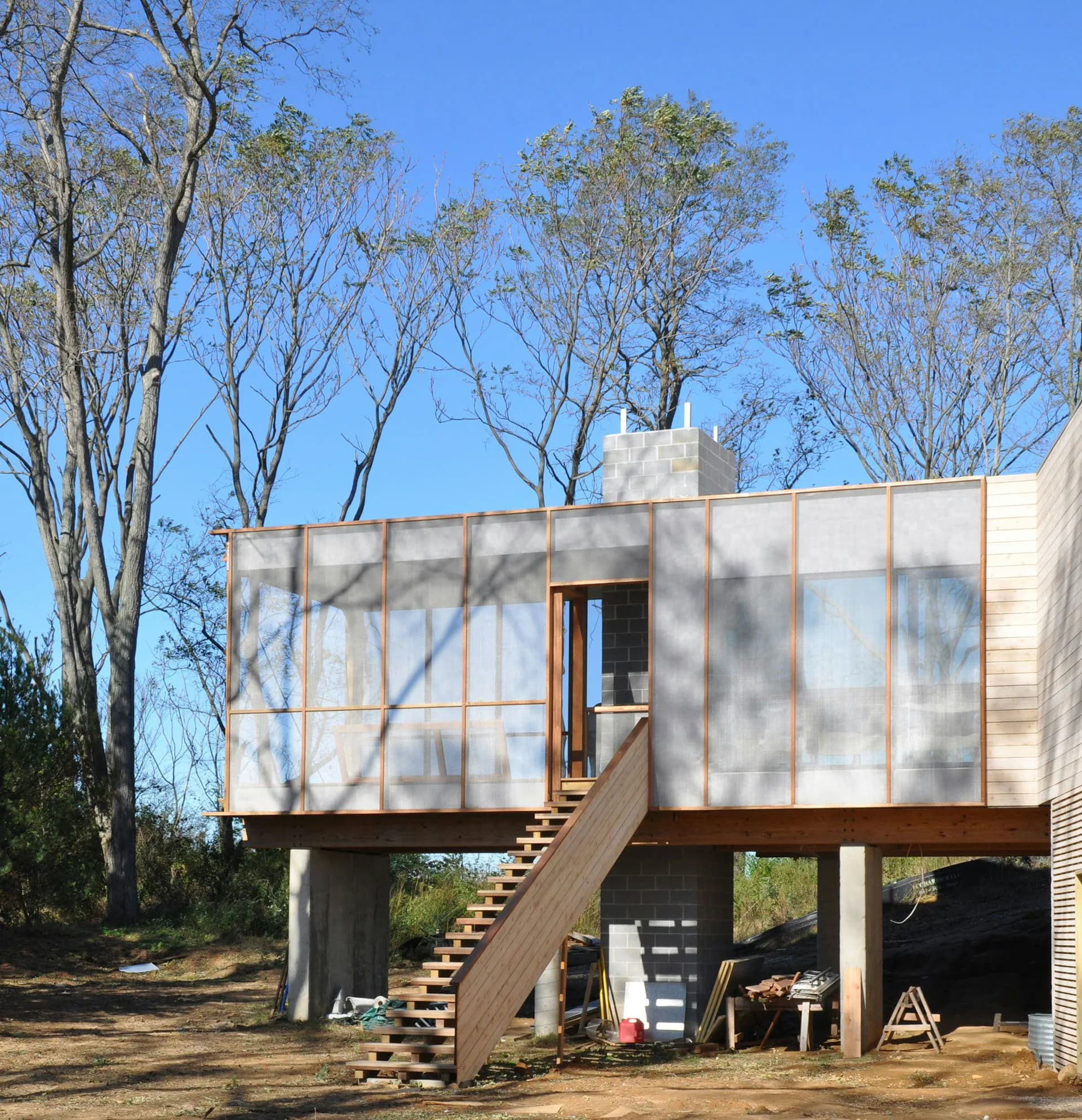 Elevated residential structure with white metal cladding and large windows, surrounded by tall trees with dappled shadows, wooden stairs leading to entrance by Ryall Sheridan Architects.