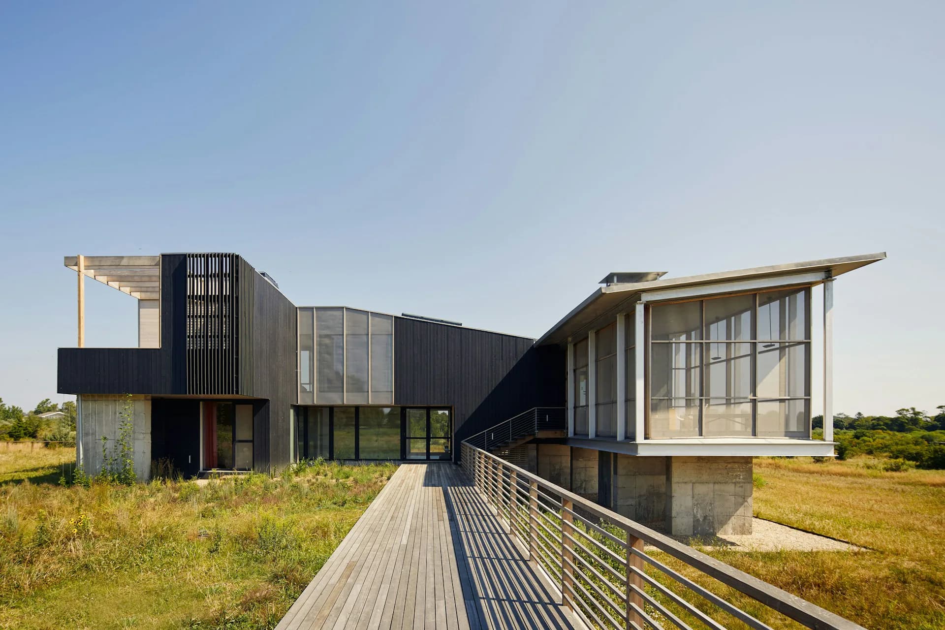 Modern residential building with black metal cladding and large glazed sections overlooking grassland, featuring a wooden deck and metal railings, designed by Ryall Sheridan Architects.