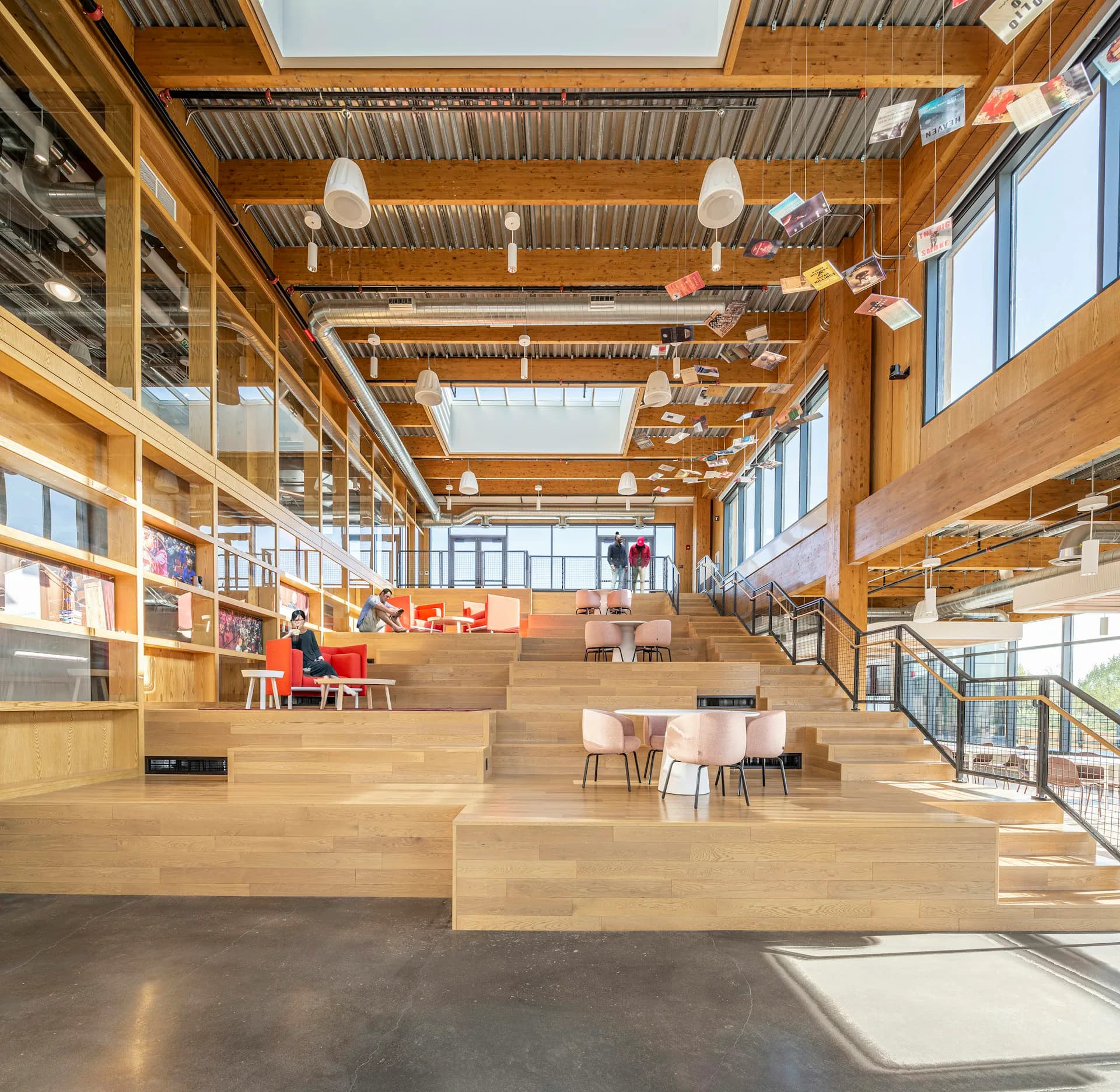 Sunlit library interior with tiered wooden platform seating, corrugated metal ceiling, large windows, and integrated shelving by S9ARCHITECTURE.
