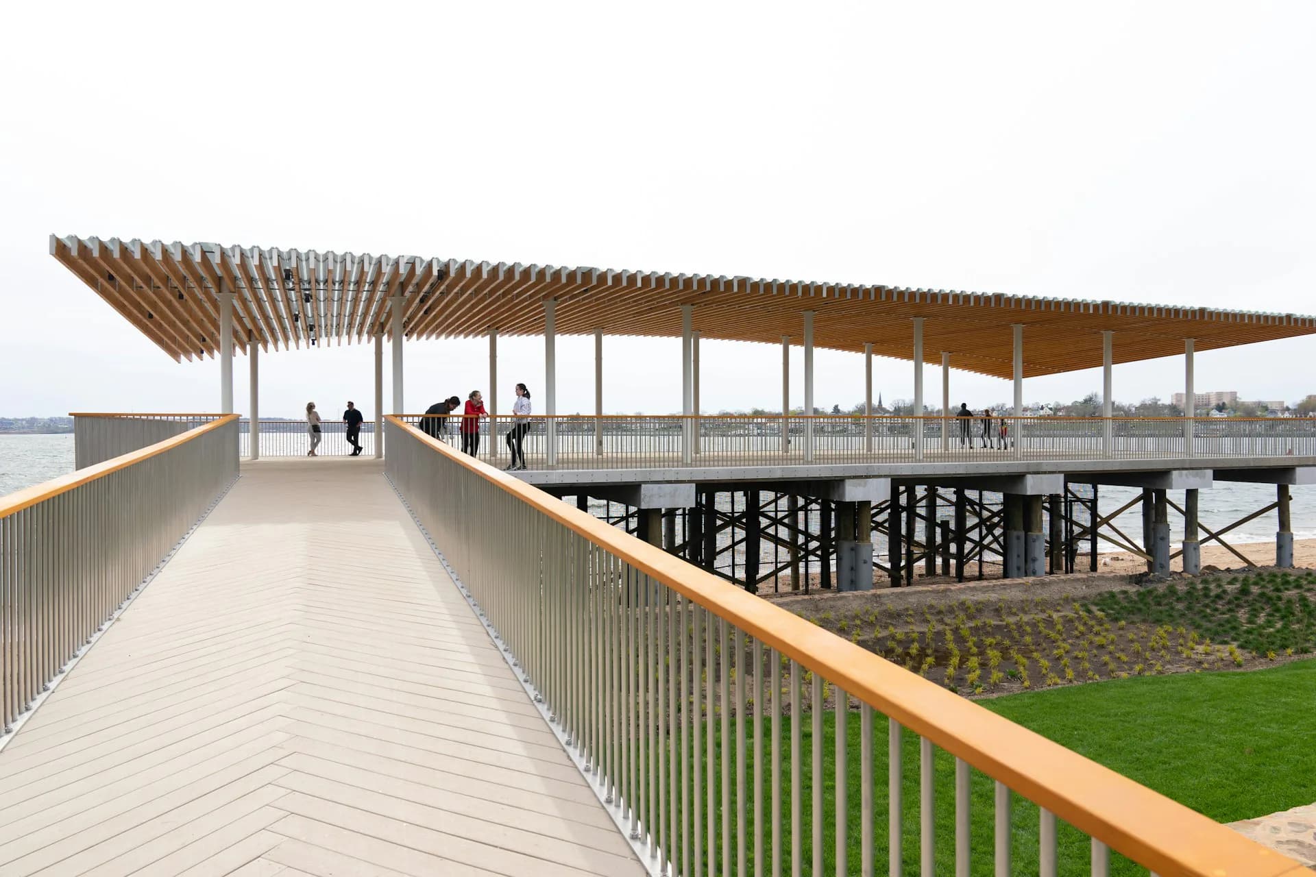Modern waterfront pavilion with wooden slatted canopy, light decking, metal railings, and integrated seating areas overlooking water and green landscape by Sage and Coombe Architects.