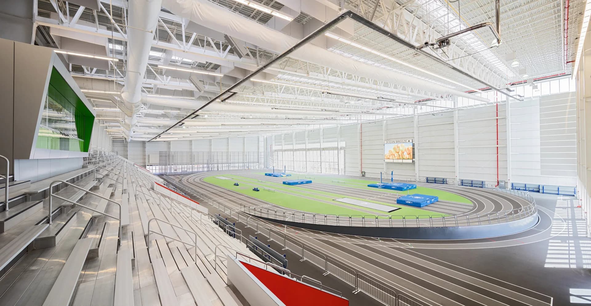 Indoor athletic facility with an oval running track in blue and green, tiered seating along one side, and industrial ceiling structure by Sage and Coombe Architects.