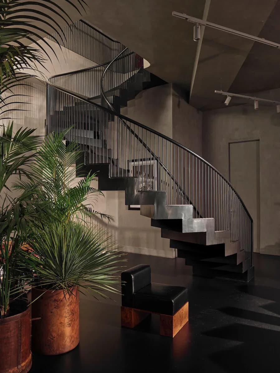 Modern interior with sculptural black and white staircase, metal railings, potted palms, and dramatic lighting by Sanayi313.