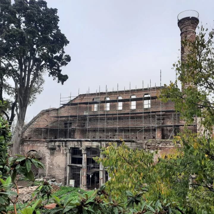 Historic brick building facade under renovation with scaffolding, ivy-covered walls, and a tall chimney, surrounded by mature trees by Sanayi313.