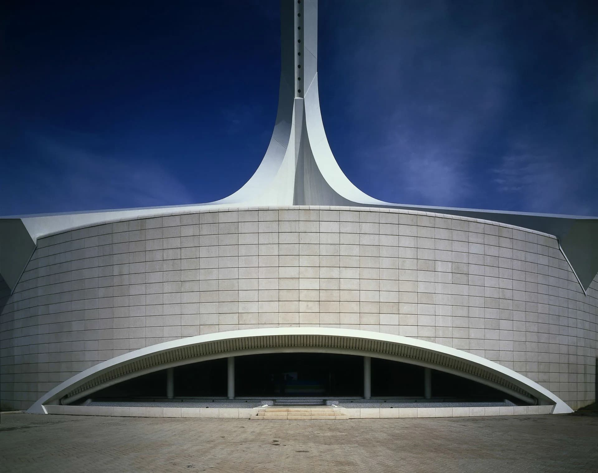 Curved white tiled building facade with dramatic sweeping arch entrance and soaring central tower against blue sky by SANTIAGO CALATRAVA.