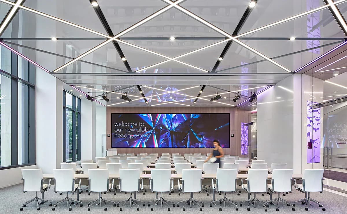 Modern conference room with geometric ceiling design, LED lighting, purple accent lighting, white chairs, and large video screen by SBLD Studio.