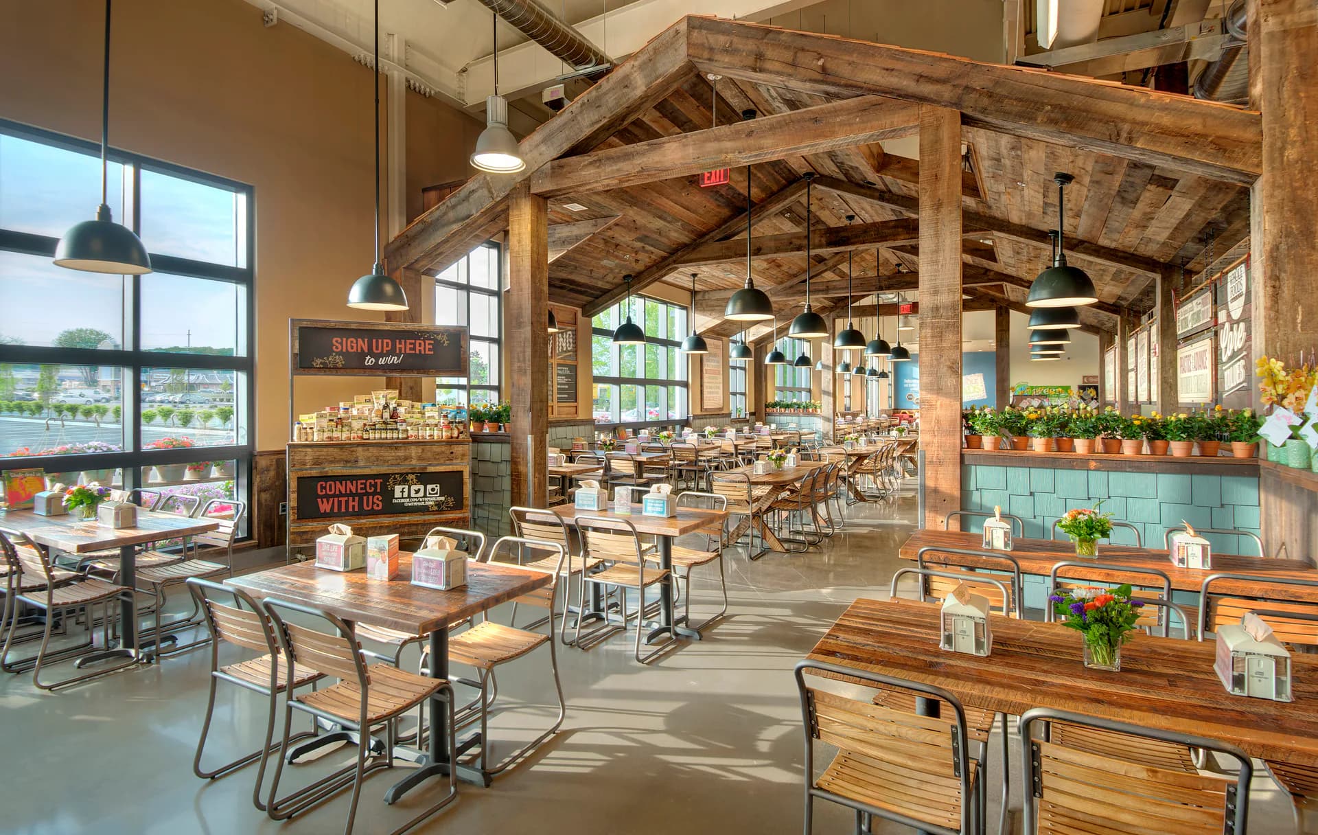 Bright industrial restaurant interior with exposed wooden beams, steel-frame tables, and large windows overlooking countryside by SBLM Architects.