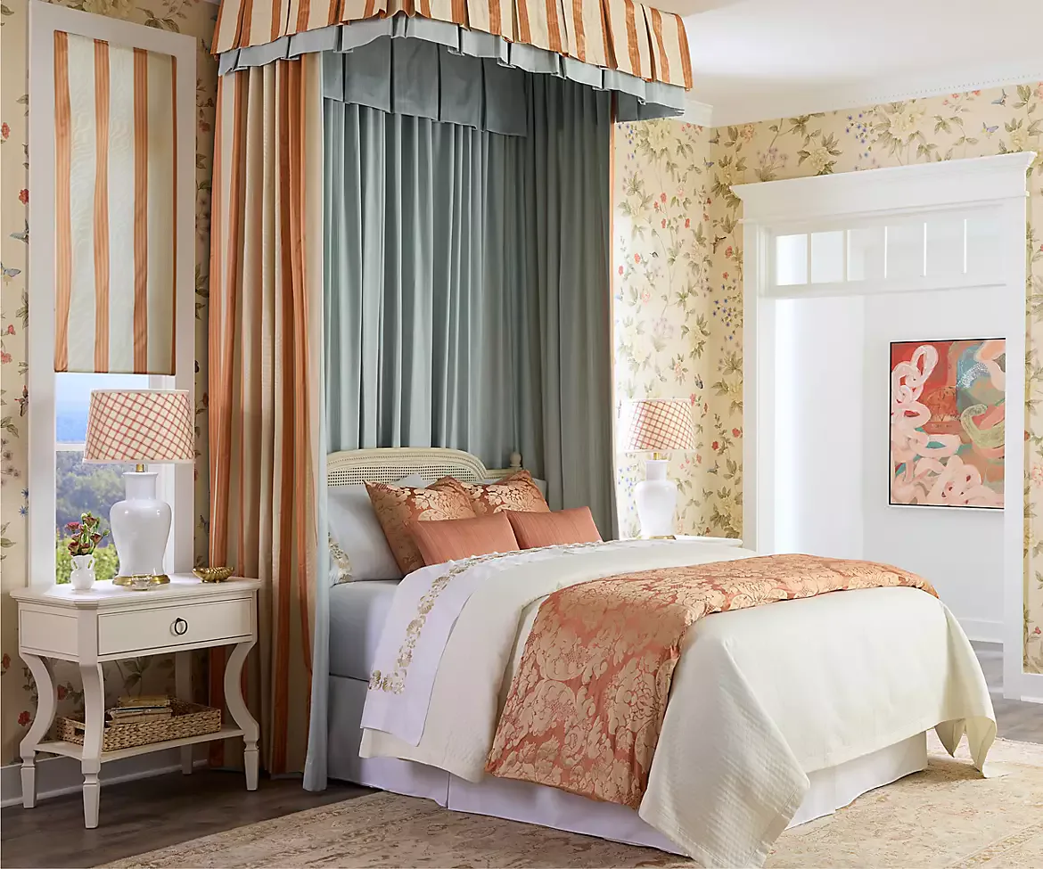 A traditionally decorated bedroom features a canopy bed with striped and floral fabrics, layered drapery in coral and gray tones, and coordinating wallpaper by Scalamandré.