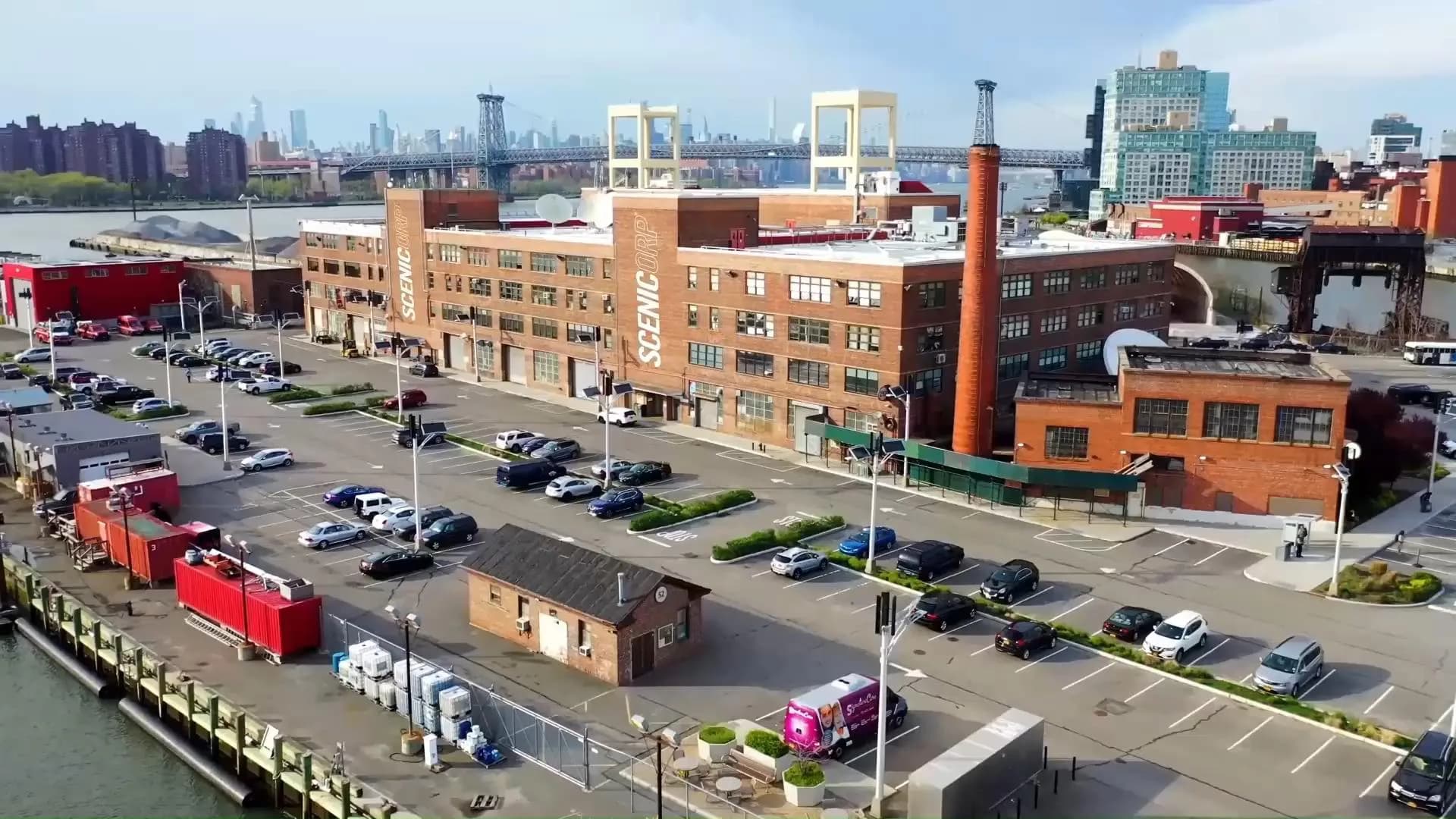 Waterfront neighborhood streetscape with brick industrial buildings, parking areas, and Manhattan skyline, landscaped public plaza by Scenicorp.